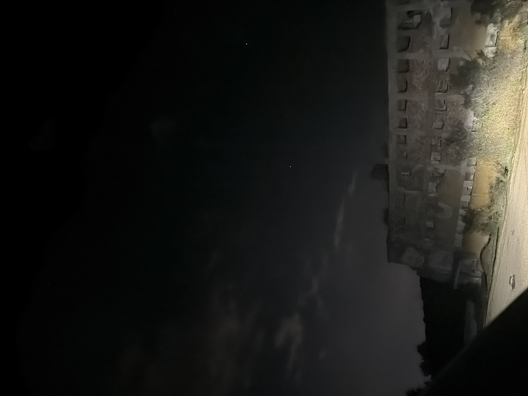 The image shows an exterior view at night, with the right side illuminated. A tall, concrete structure with regularly spaced openings is visible. Vegetation, likely grass and trees, grows beside the structure. To the left, the scene is dark, with faint reflections of the illuminated area. There are two visible stars in the night sky. The location appears to be in an urban setting, potentially in Sofia, Bulgaria. Specific details about people, subjects, and actions are not discernible.