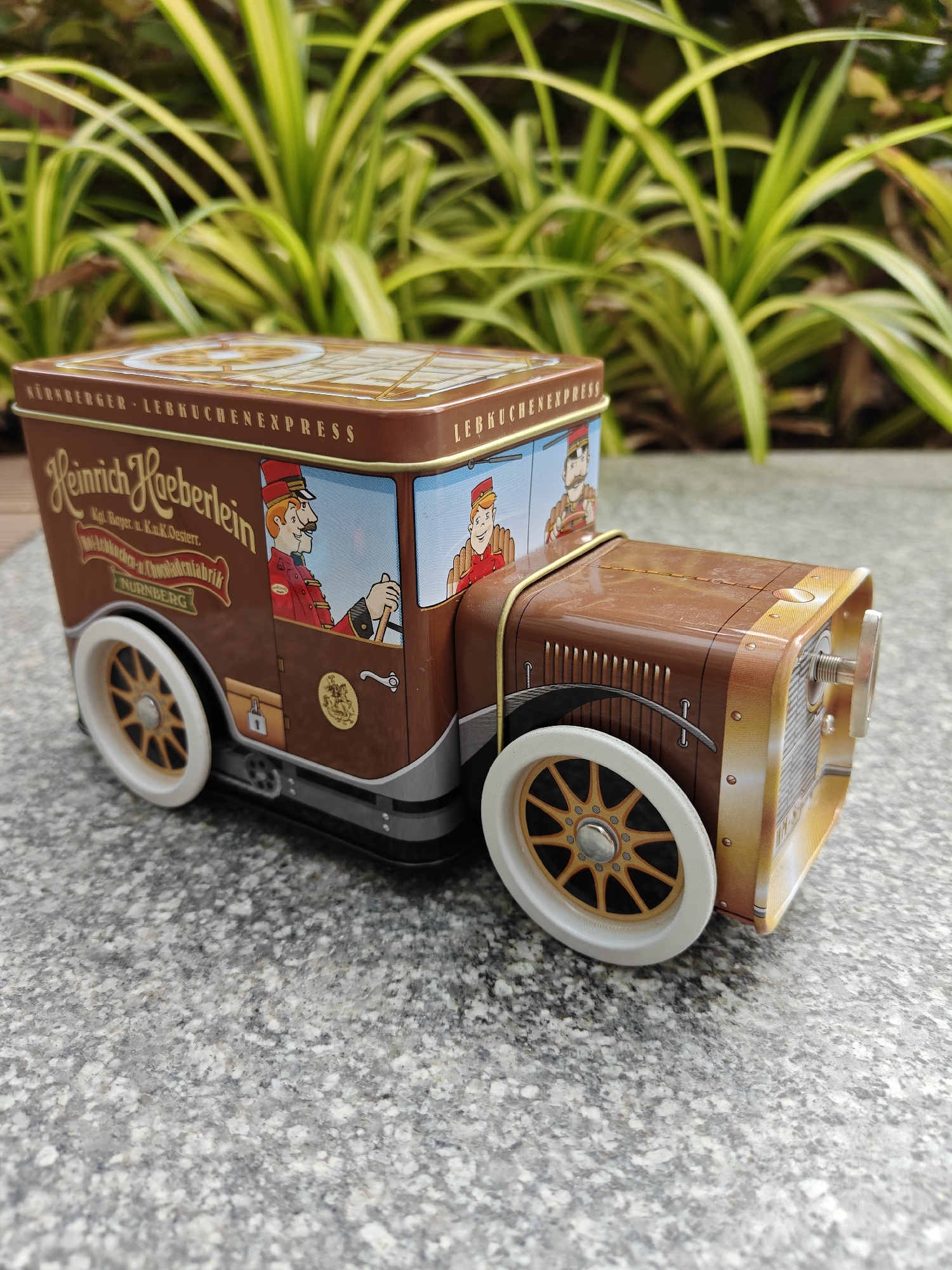 The image shows a tin box designed to resemble an antique truck. The box is brown and beige, with graphics of men in old-fashioned uniforms on the sides. The words "NÜRNBERGER LEBKUCHENEXPRESS" and "Heinrich Haeberlein" are printed on the box. The box has white wheels with gold spokes. The box is resting on a gray stone surface outdoors. In the background are green plants. There is no indication in the image that it is located in Mumbai, India.