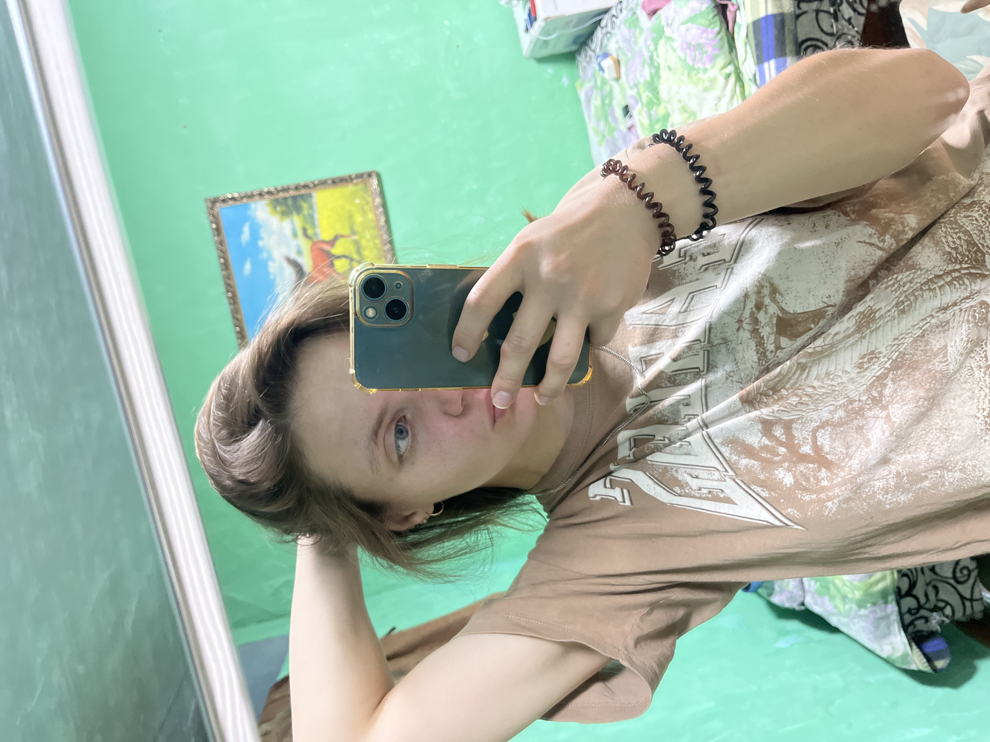 The image shows a young woman taking a mirror selfie in what appears to be a bedroom. She is wearing a light brown t-shirt with a graphic print and has her hair up. She is holding a dark green smartphone in her right hand, which is angled to capture her reflection. The background includes a bright green wall, a framed picture of a horse in a field, and a bed with patterned bedding. There are also some items on a surface to the right, possibly a box and other personal belongings. The lighting suggests an indoor setting during the daytime. This scene does not contain enough information to determine its location within the city of Nizhny Tagil, Russia.