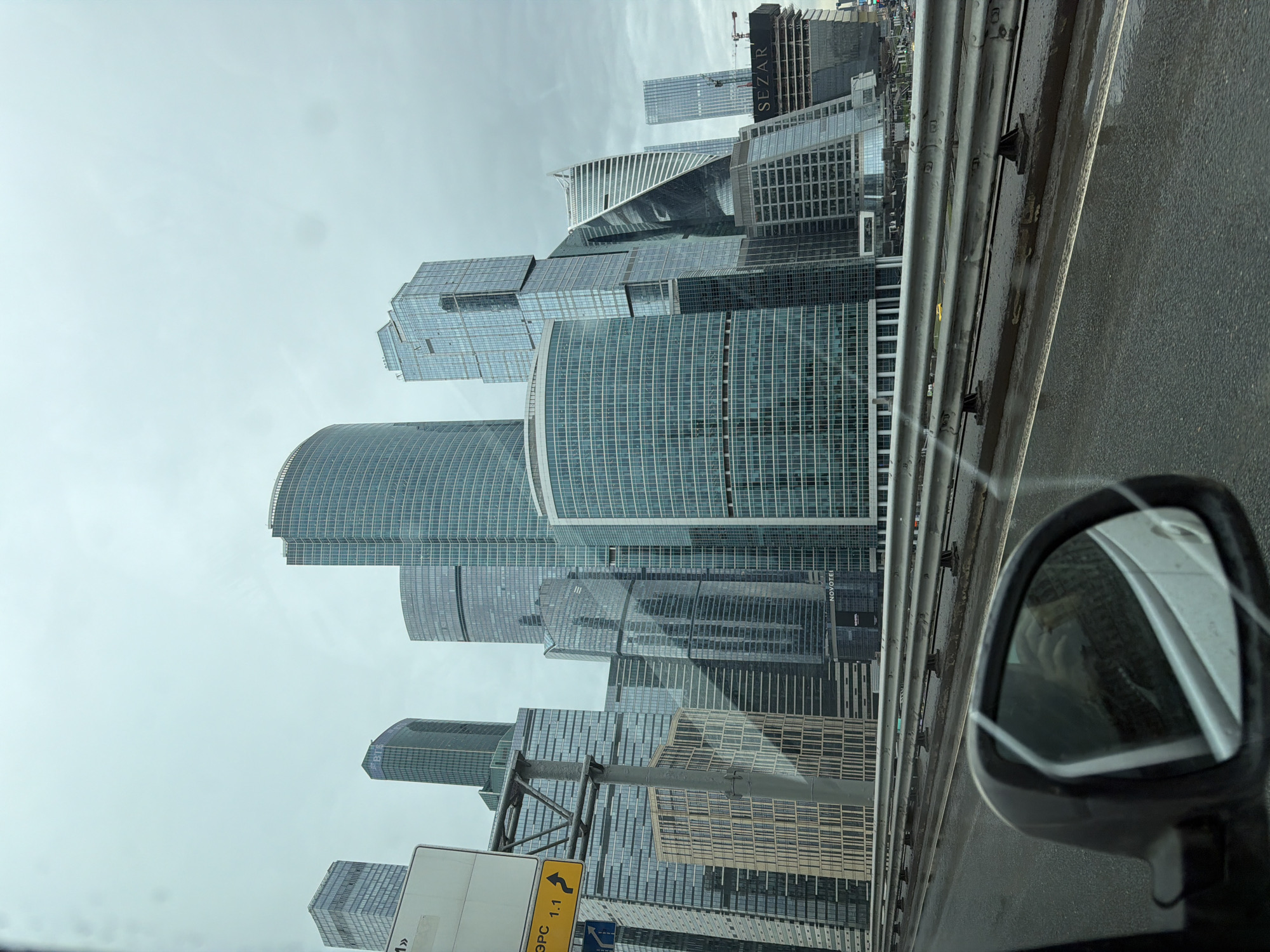 The image shows a view of Moscow International Business Center ("Moscow City") from a vehicle. Numerous modern skyscrapers with glass facades dominate the skyline, set against a gray, overcast sky. A road barrier runs horizontally across the middle of the image, with a glimpse of asphalt. The side mirror of the car is visible in the lower right corner. A street sign is partially visible on the left side of the image.
