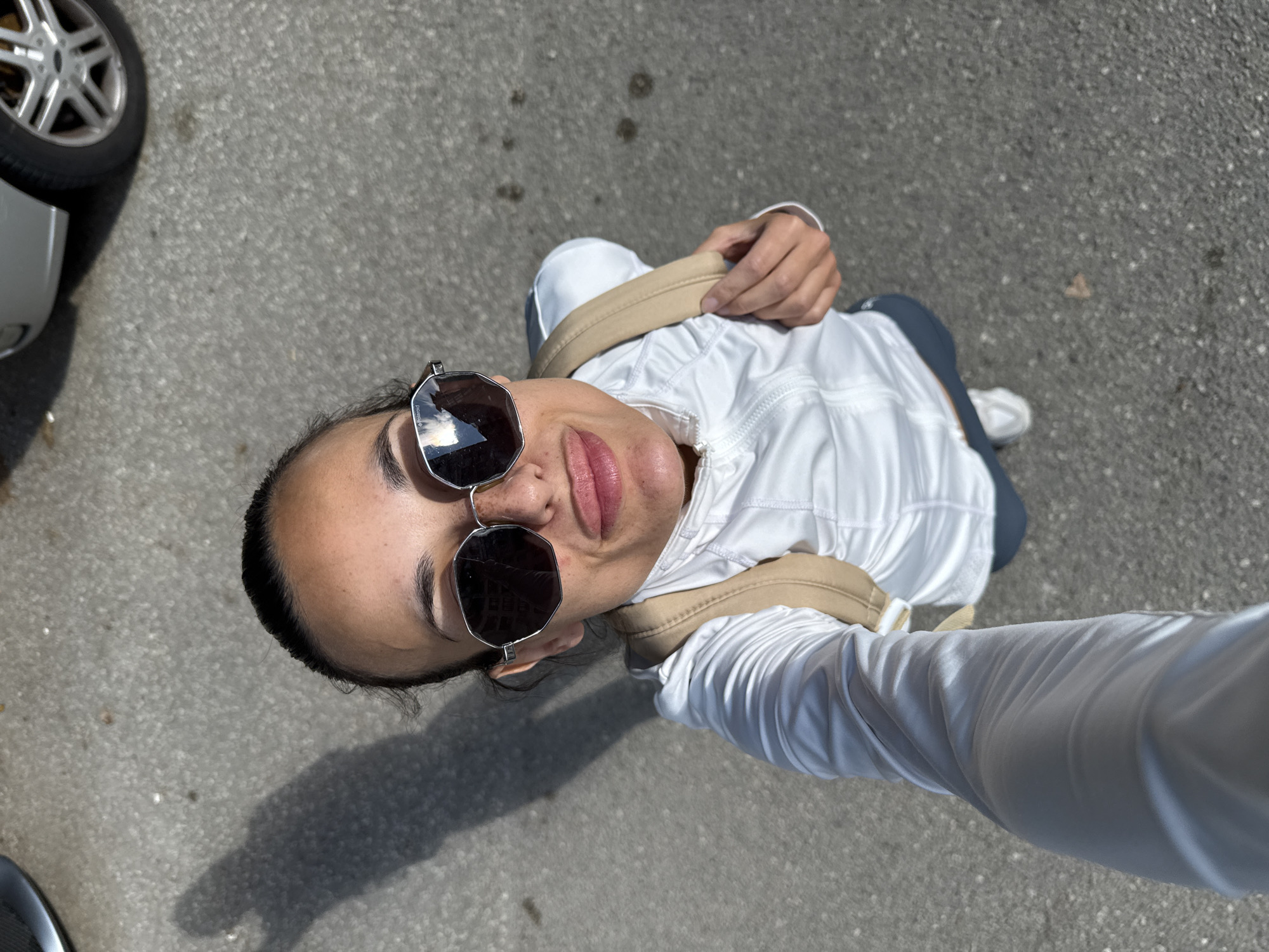 A high-angle, full-body selfie taken from the perspective of a woman standing on an asphalt surface, possibly a street or parking lot, in Pleven, Bulgaria. The woman is wearing large, octagonal sunglasses, a white zip-up jacket over a light-colored backpack, and dark leggings. Her hair is pulled back, and she is making a playful facial expression with her lips slightly puckered and her tongue visible. The ground is grey asphalt with some scattered debris and oil stains. A shadow of the woman is cast on the ground to her left. A portion of a silver car and its wheel is visible in the upper right background.