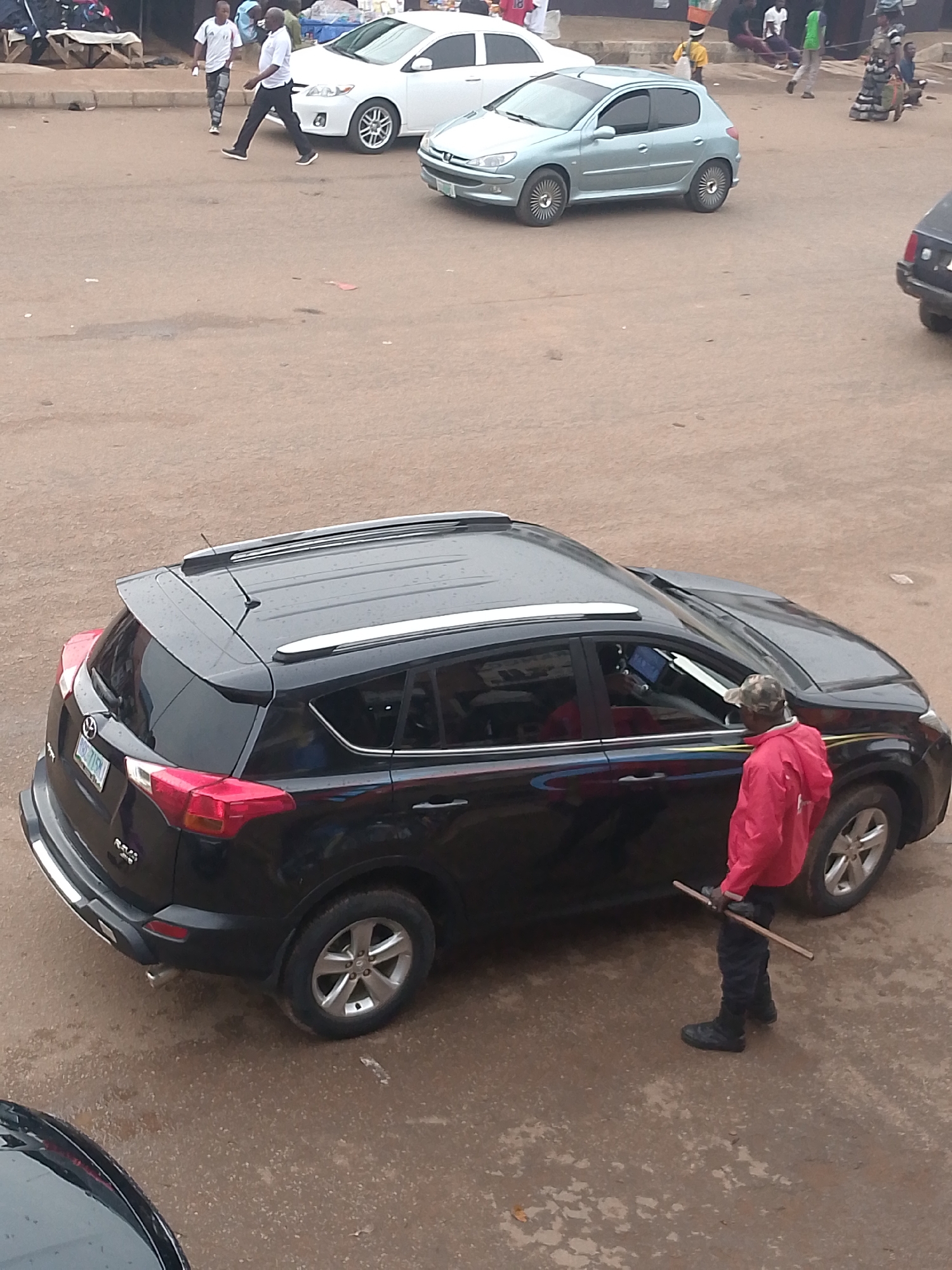 A slightly high-angle shot shows a street scene, apparently in Bukuru, Nigeria, with a black SUV parked on a dusty-colored surface. An individual in a red jacket, dark pants, and black boots stands next to the vehicle, holding a long wooden object. A white Toyota and a light blue Peugeot hatchback are parked further away. Two men walk in the vicinity of the white car. Several people are visible in the background, and another car is partially visible at the lower left edge. The overall environment suggests an urban setting.