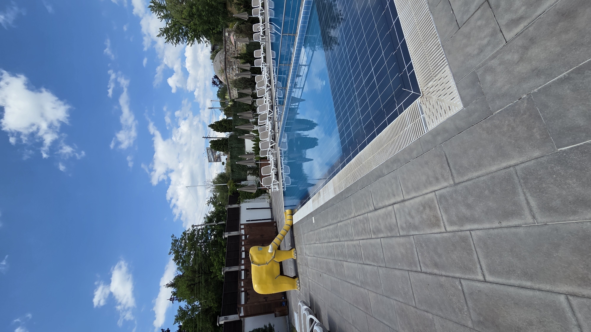 A bright yellow, cartoonish elephant statue with its trunk extended is positioned on the tiled ground next to a blue swimming pool. The pool has a tiled bottom and is surrounded by lounge chairs and umbrellas. In the background, there are trees, a fence, and some buildings. The sky is blue with scattered white clouds.
