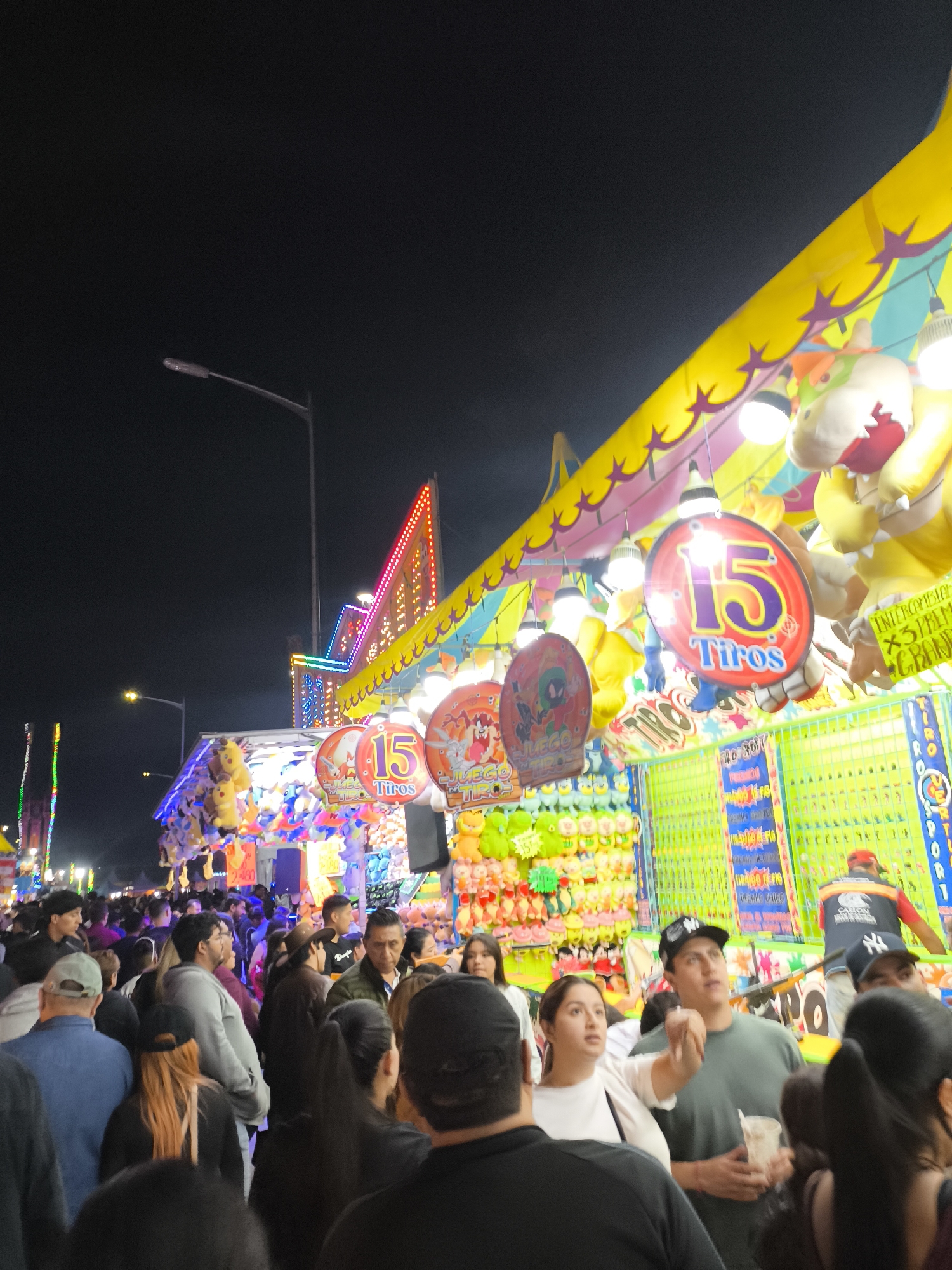 The image shows a crowded scene at what appears to be a carnival or fair, likely at night given the dark sky and artificial lighting. The location is Puebla, Mexico.
A dense crowd of people, predominantly of Hispanic ethnicity, are seen in the foreground and extending into the background. Various fair game booths are visible, adorned with bright neon lights and colorful decorations, including plush toys as prizes. The booths are labeled with "15 Tiros," suggesting a shooting or throwing game with fifteen attempts. Overhead, there are strung lights and colorful awnings. Streetlights are visible in the background. People are milling about, some looking at the booths, others seemingly conversing. A few individuals stand out: A man in a green shirt holds a cup while looking upwards. Several are wearing baseball caps. The crowd is dense and diverse in attire, suggesting a lively and festive atmosphere.