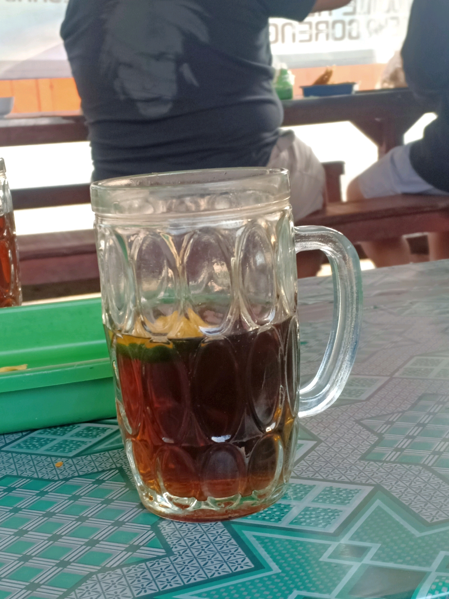 A close-up, eye-level shot shows a glass mug filled with a dark beverage, likely iced tea, with a layer of ice and possibly lemon visible at the top. The mug has a thick, ribbed glass design and a prominent handle. It sits on a patterned tablecloth with a green and white diamond design. To the left of the mug, a green plastic tray is visible. In the background, two individuals are partially seen sitting at a wooden table. The person on the left wears a dark t-shirt and white shorts. The person on the right wears a dark shirt. The environment appears to be an outdoor eating area, possibly a warung or cafe, in Jatisrono, Indonesia, suggested by the lighting and casual setting.