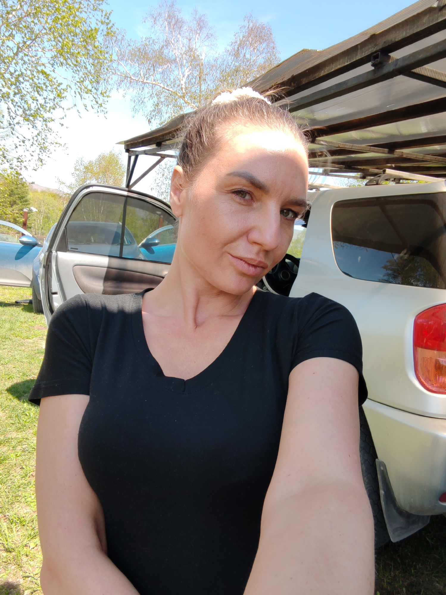 The image depicts an adult female engaged in a selfie in an outdoor setting in the city of Unknown, Unknown. The subject, positioned centrally and angled slightly to her right, has light skin, light brown hair pulled back, and green eyes. She wears a black V-neck t-shirt. A distinct, thin linear scar or crease is visible on the left side of her neck near the collarbone. Her expression is neutral with slightly pursed lips.
In the background, behind the subject, a light-colored SUV or crossover vehicle is parked on grass, with its rear right quarter visible, including a red taillight and a black mudflap. To the left of the SUV, a silver car is parked on the grass with its front passenger door open. Further back and to the left, a light blue car is also visible with an open door.
Above the vehicles, a simple canopy structure with a dark wooden frame and translucent corrugated roofing material is present. The ground is covered in green grass. Numerous trees with sparse green foliage and some bare branches are visible against a clear, bright blue sky. The scene is illuminated by strong daylight, casting shadows.