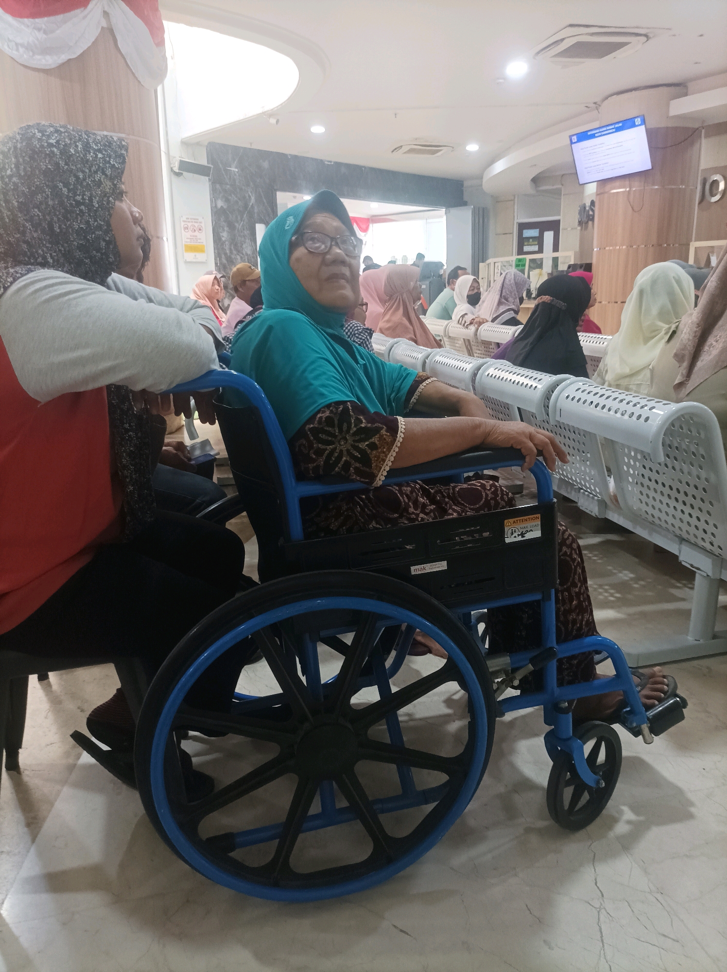 The image shows an indoor waiting area, likely a hospital or clinic, in Sumuragung, Indonesia. In the foreground, a woman wearing glasses and a teal headscarf is seated in a blue wheelchair. She is dressed in a patterned brown and teal outfit. To her left, another person wearing a grey top and a dark patterned headscarf is partially visible, leaning on the armrest of the wheelchair. Behind them, a row of white metal benches is occupied by several other individuals, mostly women wearing headscarves. Some are looking towards a screen displaying information on the wall in the background. The floor appears to be tiled. The overall atmosphere suggests a calm waiting environment.