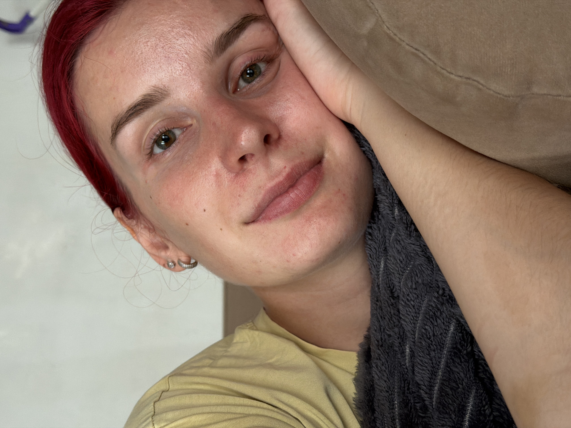A young woman with bright red hair is lying on her side, resting her head on her hand and arm, which is partially obscured by a dark gray blanket. She is looking towards the camera with a neutral expression. She is wearing a yellow t-shirt. The background consists of a light-colored wall and a brown sofa. The scene appears to be indoors. There is no indication of the location being in the city of Chaikovsky, Russia.