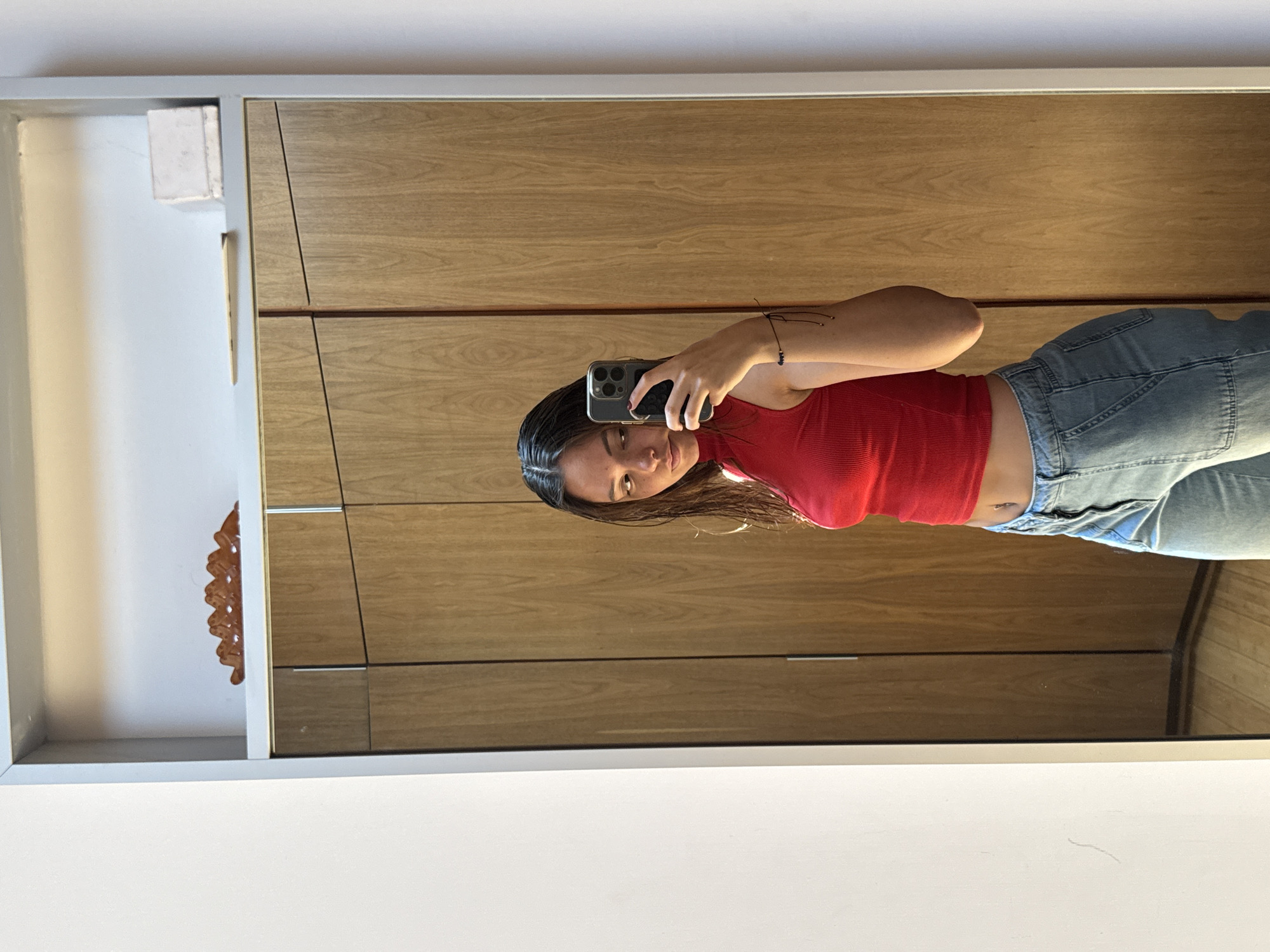 A woman is taking a selfie in a mirror. She is wearing a red top and light-colored jeans. She is holding a smartphone in her hand, and her face is visible in the reflection. The background includes a light wooden wardrobe and white walls. There is a decorative object partially visible in a niche to the left. The location is a residential setting, likely indoors. Without additional information, pinpointing the precise location in San Bernardino Tlaxcalancingo, Mexico, is impossible.