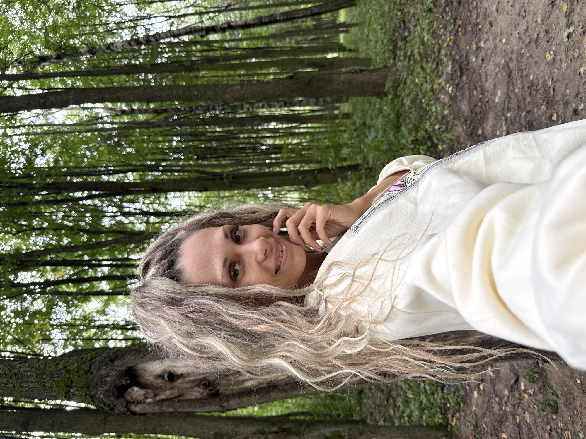 A woman with long, wavy blonde hair, some streaks of pink, is in the center of the frame, smiling at the camera. She is wearing a cream-colored sweatshirt. Her left hand is near her face, touching her chin. The background is a forest with many tall, slender trees and green foliage. The ground is covered with dirt and fallen leaves. The lighting suggests it is daytime, and the scene is likely outdoors in a natural setting. The image appears to be a selfie. There is no identifiable indication of the location being in Rusanovo, Russia, or any other specific city or country.