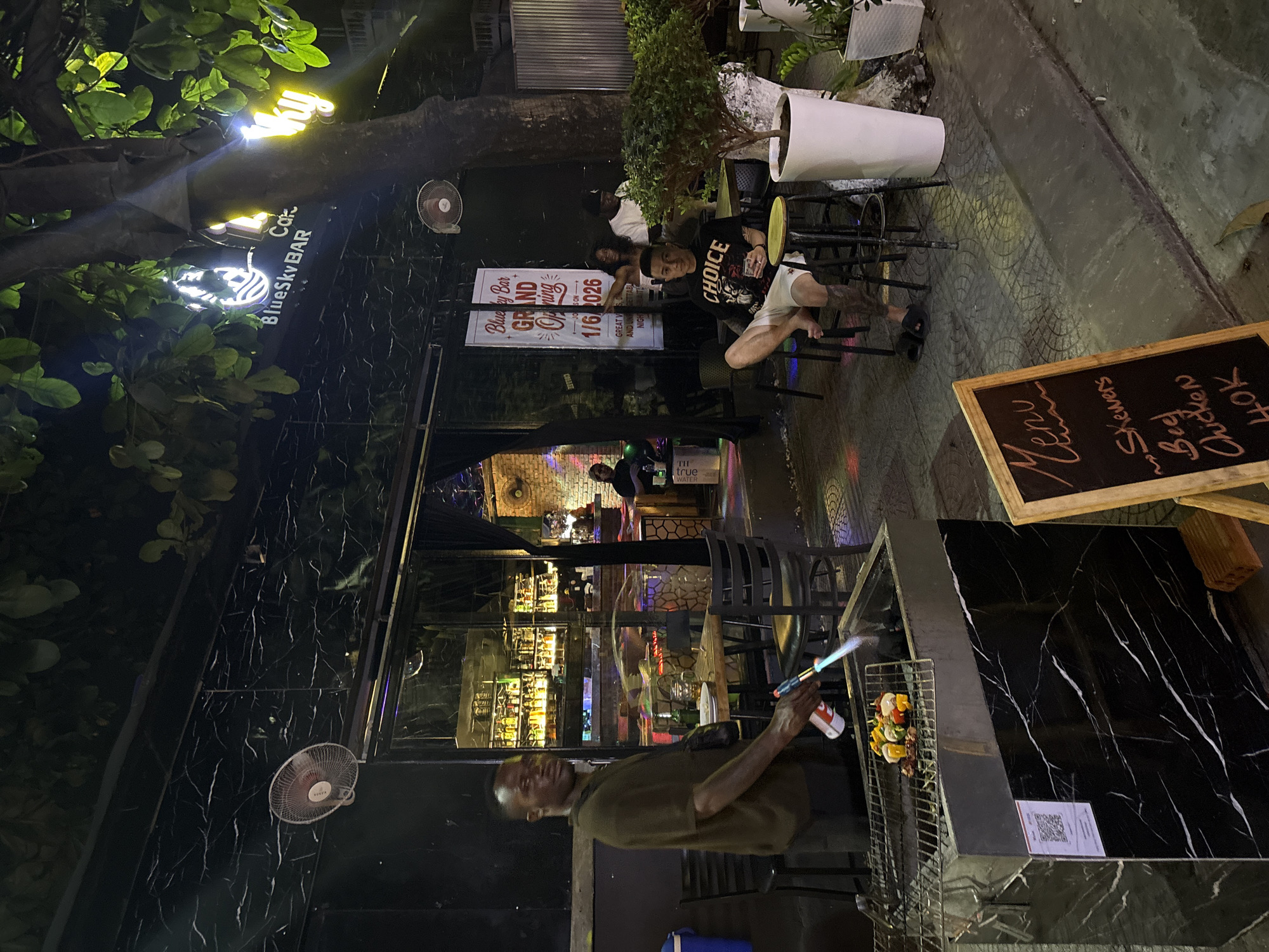 Here's a detailed description of the image:
**Setting:** The image appears to be taken at night in what looks like an outdoor seating area connected to a bar. The location is likely in Đà Nẵng, Vietnam. This assumption is based on the bar's name, "BlueSky BAR," and its grand opening date of 1/6/2026, which follows a common date format used in that region. The surroundings are urban, with marble-patterned walls.
**People:**
* **Grilling Person:** A man stands near a grilling station. He is using a small torch to cook skewers of meat and vegetables. He's wearing a green-brown shirt.
* **Customers:** A group of customers are seated in the outdoor area. One is wearing a black t-shirt with the word "CHOICE" on it, and white shorts, and appears to be barefooted.
**Objects:**
* **Bar/Restaurant Signage:** There's a sign advertising the "BlueSky BAR" grand opening on "1/6/2026".
* **Menu Board:** A menu board displays handwritten options such as "Beef Skewers" and "Chicken", with prices.
* **Grilling Station:** A stainless steel grill with skewers being cooked.
* **Tables and Chairs:** Outdoor seating is arranged.
* **Fans:** Several fans are mounted on the wall.
* **Lights:** There are decorative lights and trees visible.
* **Plants:** Several plants are visible, some in white pots.
**Actions/Interactions:**
* **Cooking:** The man is actively grilling skewers.
* **Customer Interaction:** Customers are seated and appear to be enjoying their drinks.
**Environment:**
* The scene is at night, with artificial lighting illuminating the area.
* The walls adjacent to the bar is covered in a marble pattern.
* The general atmosphere seems to be a lively and casual outdoor bar setting.