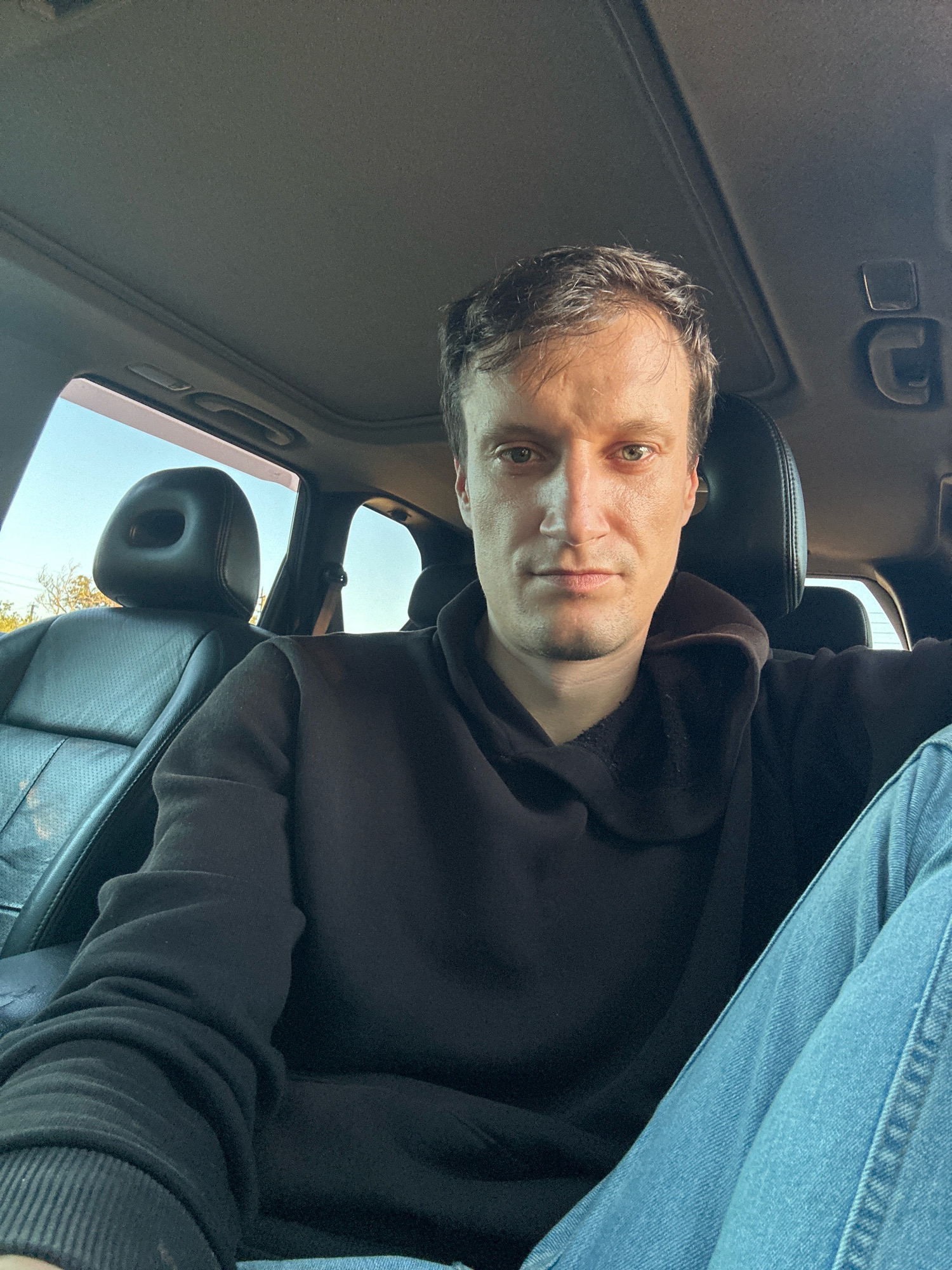A man with short brown hair and green eyes is sitting in the driver's seat of a car, looking directly at the camera. He is wearing a black hooded sweatshirt and blue jeans. The interior of the car is visible, including the black leather seats and the sunroof. The background outside the car shows a bright sky and some trees. The image appears to be taken during the day.