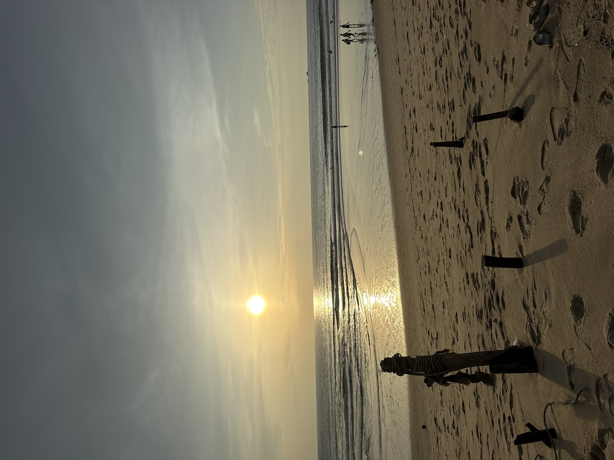 The image depicts a beach scene at sunset in Choeng Thale, Thailand. The sun is low on the horizon, casting a warm glow across the sky and reflecting brightly on the water. Gentle waves roll towards the shore, leaving a wet, sandy expanse that mirrors the sky. In the foreground, several weathered wooden posts, some wrapped with rope, emerge from the sand. Footprints are visible across the beach, suggesting recent activity. In the middle ground, a few figures are walking along the shoreline, appearing as silhouettes against the bright water. Further out in the ocean, there are additional small figures and what appears to be a distant boat on the horizon. The sky is a gradient of pale yellow near the horizon transitioning to a muted gray-blue overhead, with wispy clouds scattered throughout. The overall impression is one of tranquility and the end of a day at the coast.