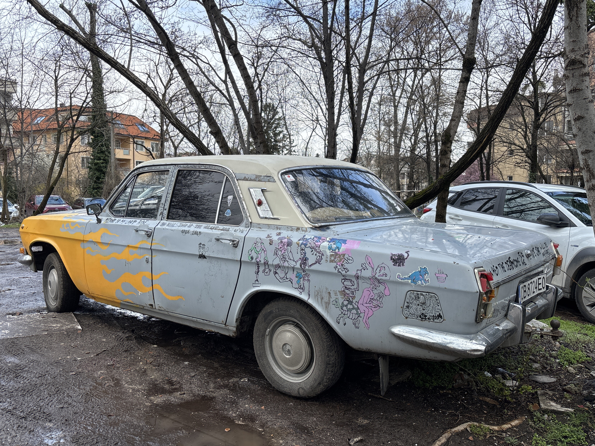 An old, grey, and beat-up Volga car with a yellow front fender and door stands in a muddy, tree-lined area. The car is adorned with various stickers and drawings. The license plate reads "CB 8724 EO". Behind the Volga is a white SUV. Bare trees surround the scene. In the background, a building with a red tile roof is visible. The location is likely Sofia, Bulgaria, based on the license plate format. The ground is covered in mud and leaves.
