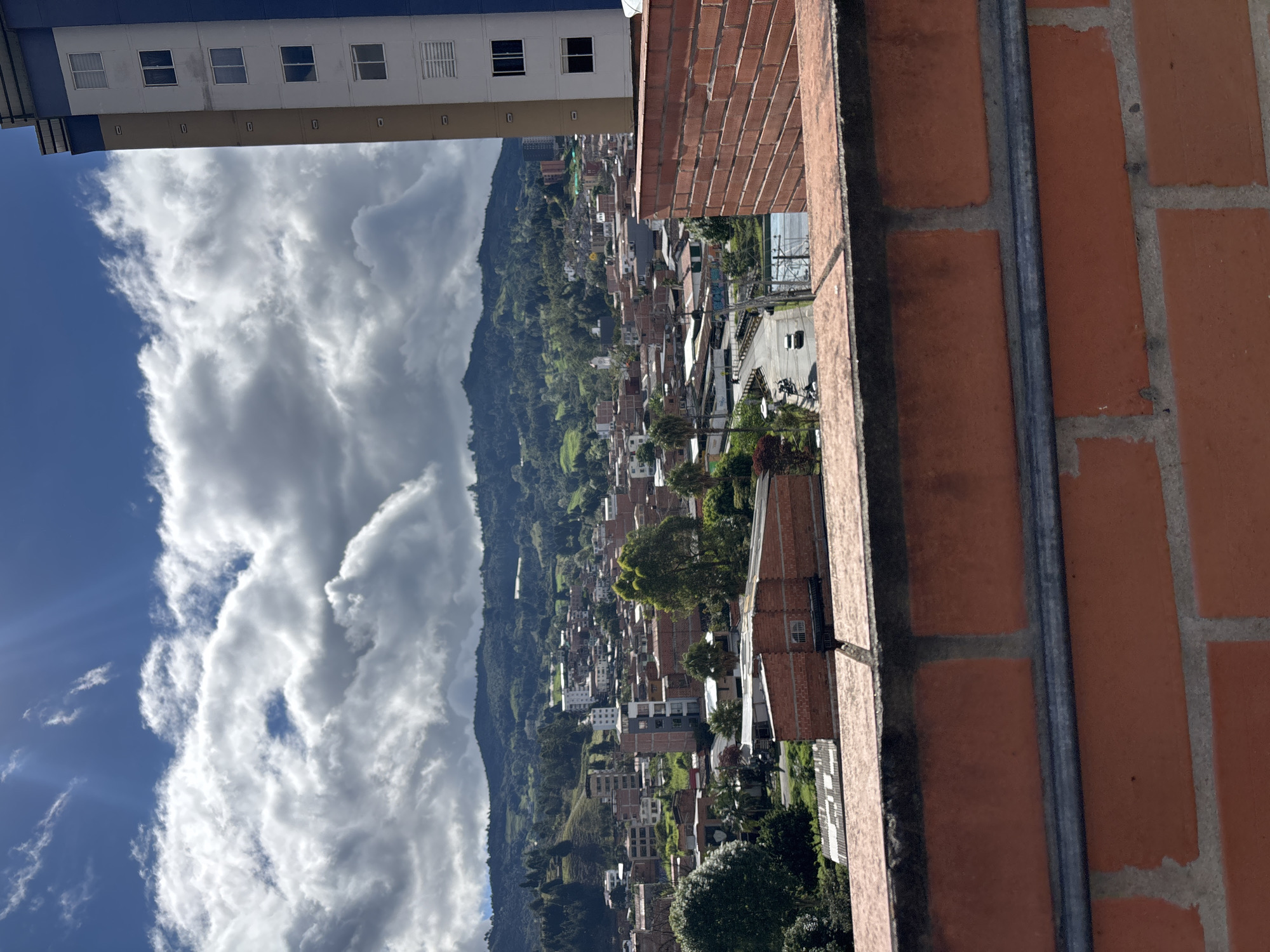 The image depicts a view from a building in Guarne, Colombia. A brick wall is in the right foreground. Through an opening, the view shows a valley with numerous buildings scattered across the landscape. A mountain rises on the left side of the valley, with clouds partially obscuring it and the blue sky. The top left corner shows the edge of another building.