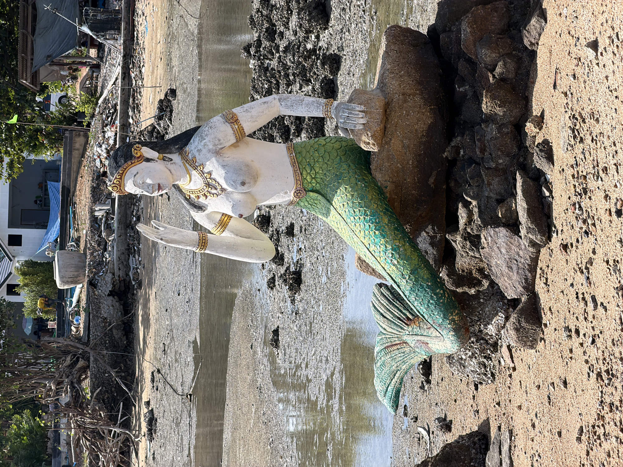 A statue of a mermaid sits on rocks at the edge of a body of water, possibly the sea, in Baan Plai Laem, Thailand. The mermaid statue is depicted with a human upper body and a green, scaly fish tail. She is adorned with jewelry, including a crown, necklaces, and bracelets. The statue is white with painted gold accents and black hair.
The environment surrounding the statue is a beach with sand and rocks. In the background, there are signs of human activity and possibly some disarray. There are structures that appear to be buildings, some with roofs. Debris is visible on the shore and near the water, including what looks like discarded materials and possibly building remnants. Trees and vegetation are present in the background. The water appears somewhat murky and shallow, with exposed patches of sand and mud. The lighting suggests it is daytime.