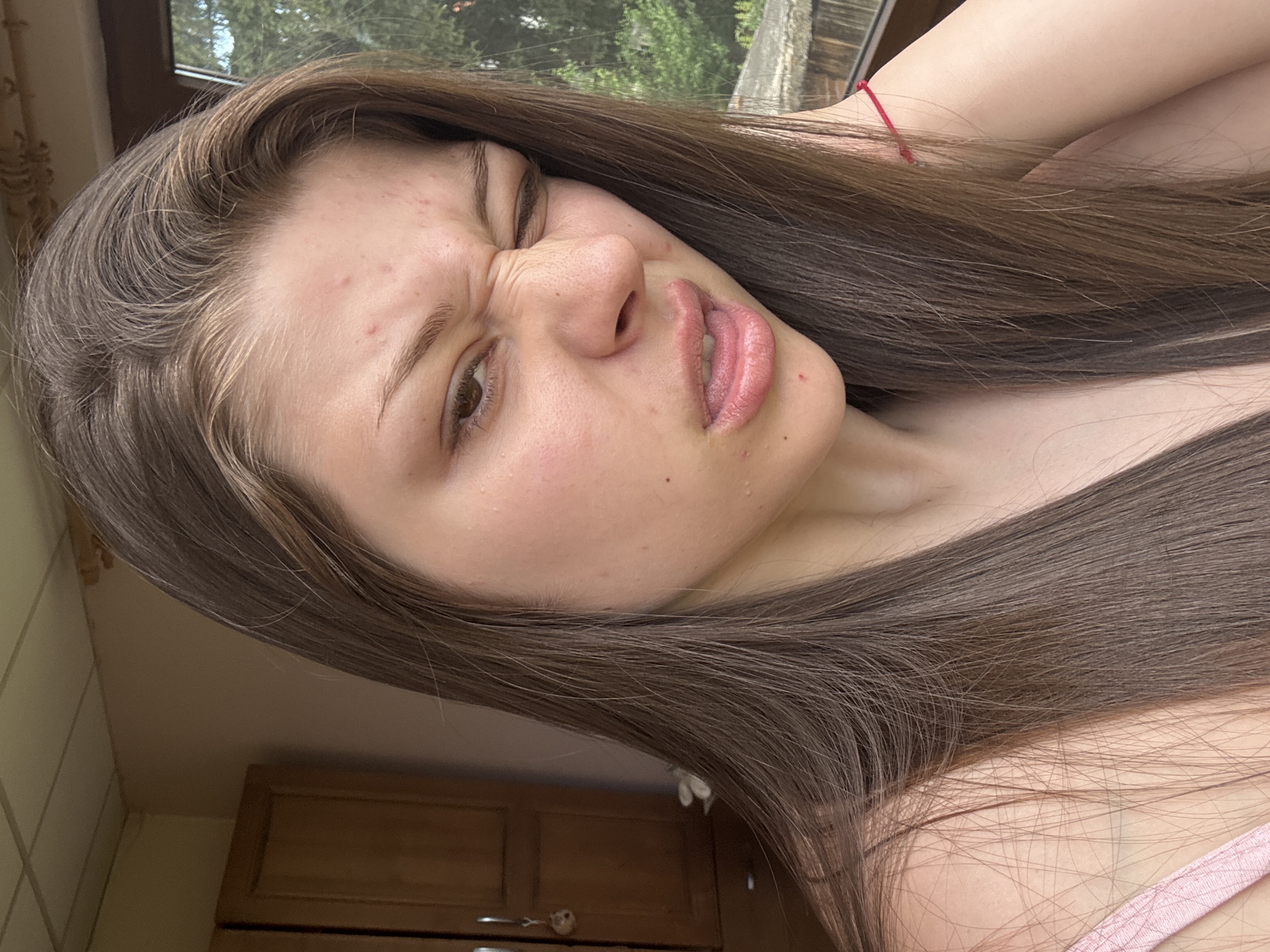A close-up, vertically oriented, low-angle shot shows the face of a young person with long, dark brown hair. The individual is looking towards the viewer with their eyes squinted and their mouth contorted in a playful or exaggerated expression, with their lips slightly parted to reveal their teeth. Their eyebrows are furrowed, and there are visible pores and some blemishes on their skin, particularly on the forehead. The lighting suggests it is daytime, possibly outdoors or near a window, as indicated by the soft, natural light illuminating their face and the hint of greenery and a window frame in the background. The person is wearing a pink strap, likely from a garment, visible on the right side of the frame, and a thin red string is tied around their wrist. The background is out of focus, featuring a wooden cabinet and wall, suggesting an indoor setting with a window view. The overall mood is lighthearted and casual, possibly a selfie taken in a relaxed environment. No visible text is present in the image.