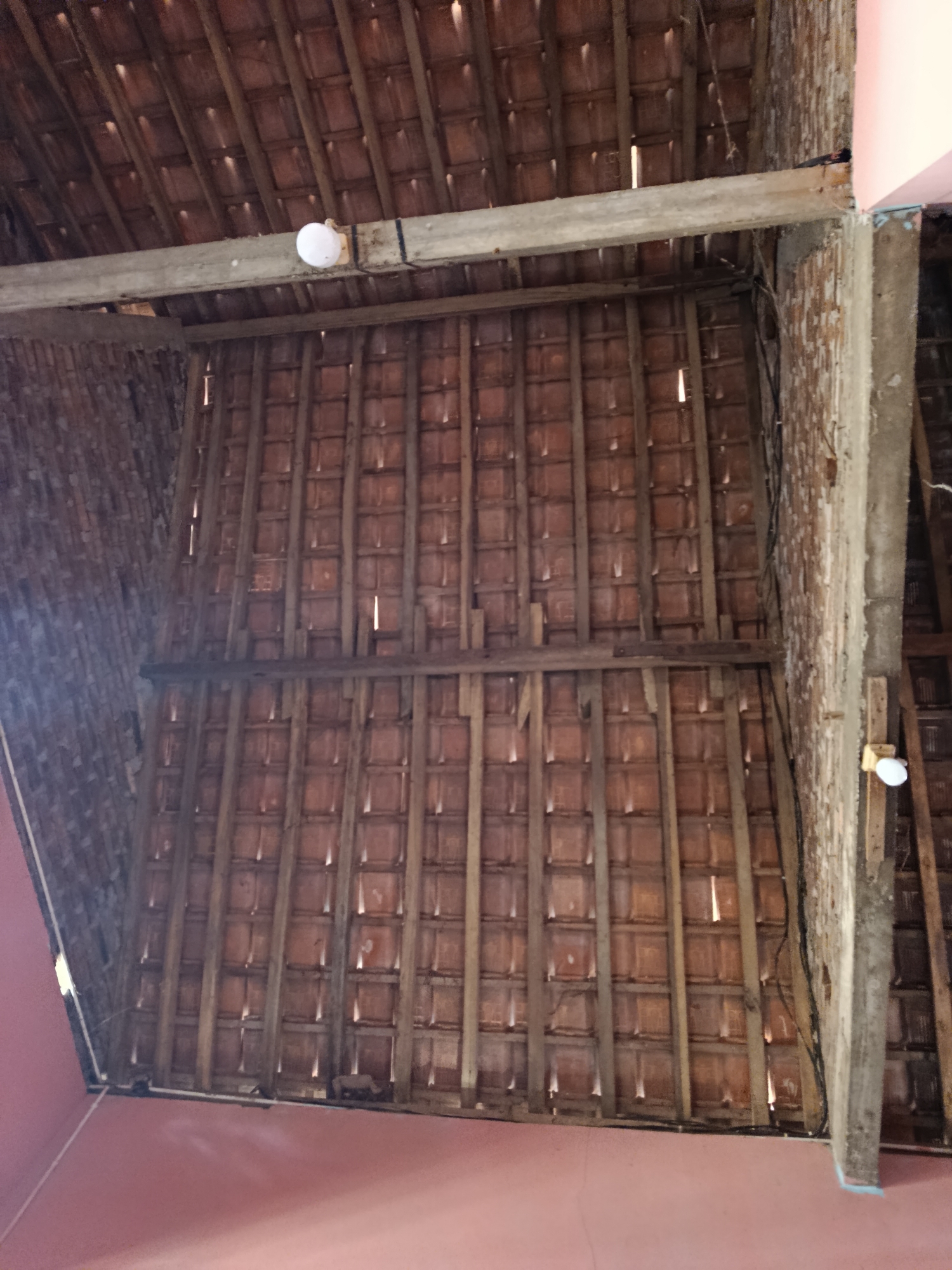 The image captures the underside of a roof constructed with terracotta tiles and exposed wooden beams. The roof structure is supported by brick walls, one of which is visible on the right side of the image. Several electrical wires run along the roof and down the wall. Two simple light fixtures are visible, one mounted on a horizontal beam near the center-left and another on a vertical wooden post near the bottom right. The overall impression is of a partially constructed or rustic interior space, possibly a barn or an unfinished building. The location is identified as Sedayu, Indonesia.