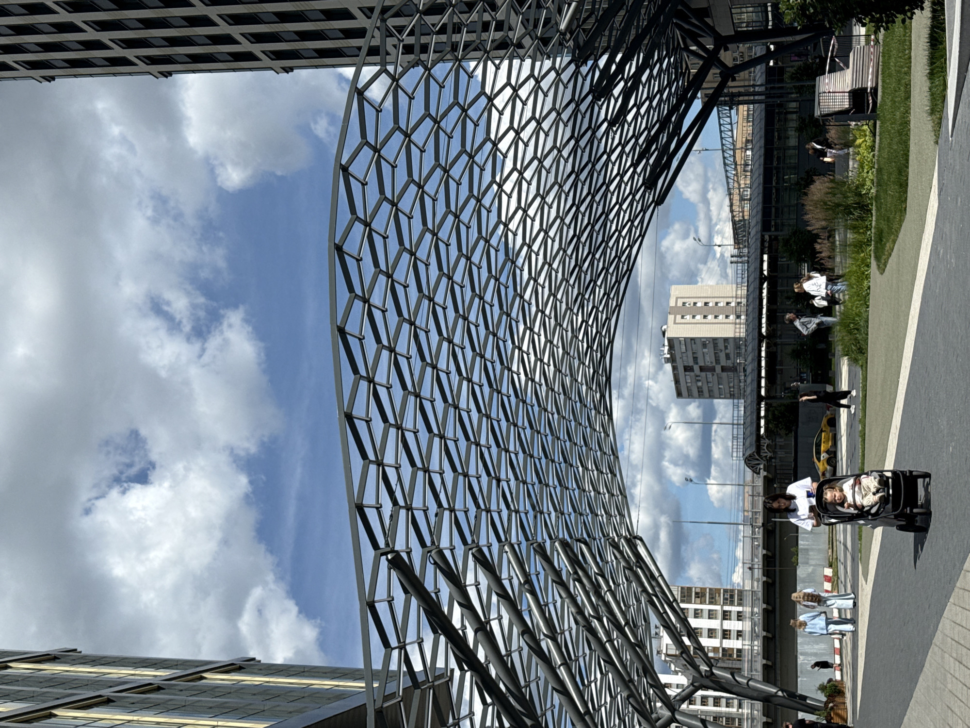 The media file shows an outdoor scene in Moscow, Russia, on a bright, cloudy day. The foreground is dominated by a modern, geometric metal structure with a hexagonal pattern, possibly a canopy or part of a building facade. This structure curves upwards and to the right, with a sky filled with white, fluffy clouds against a blue backdrop visible through its openings.
To the right of the structure, a park-like area with green grass and a paved walkway is visible. Several people are present in this area. In the middle ground, a woman in white is pushing a stroller with a child inside. Further in the distance, other individuals are seen walking or sitting. A yellow car is parked in the background.
Behind the park area, buildings are visible, including a multi-story structure with many windows. The overall impression is one of a contemporary urban environment with recreational spaces. The lighting suggests it is daytime, likely midday or early afternoon. The scene appears calm and everyday, with no specific event clearly depicted, rather a snapshot of life in a public space. No visible text is present in the media file.