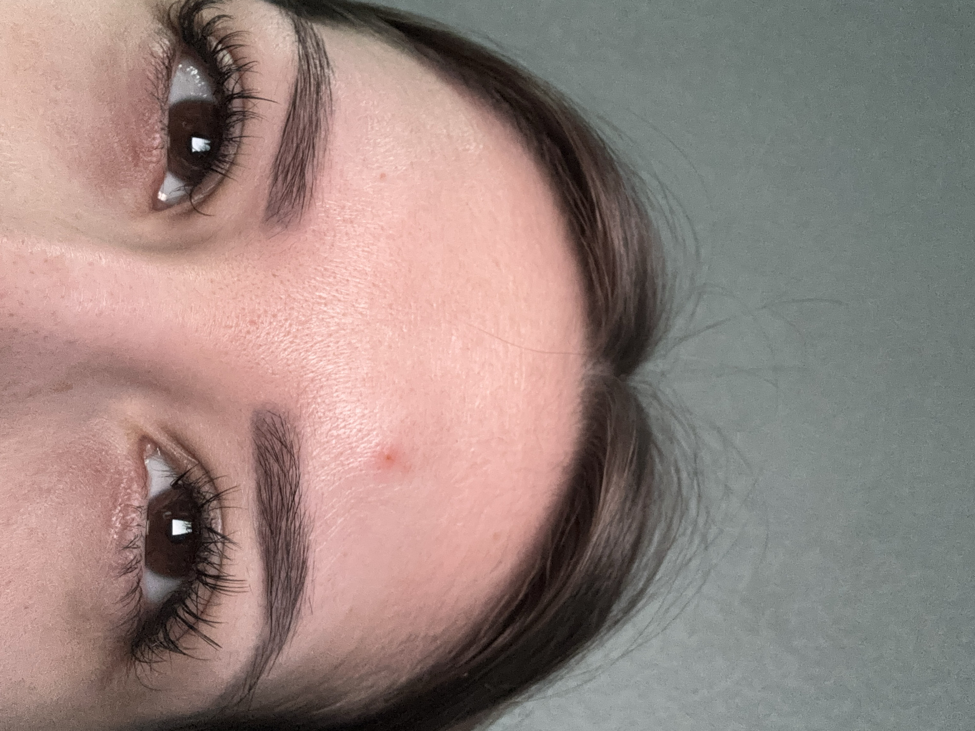 The media file is a close-up, rotated image of a person's face, focusing on their eyes, eyebrows, and forehead. The subject has dark brown eyes with long, dark eyelashes and well-defined, dark eyebrows. Their skin appears fair with some visible pores and a small reddish blemish on the forehead. A strand of dark brown hair is visible on the right side of the frame. The background is a plain, out-of-focus gray surface. The lighting suggests an indoor setting, possibly with artificial light, as there are reflections visible in the eyes. The image is cropped tightly, with only a portion of the face visible, and it is rotated approximately 90 degrees counter-clockwise. No specific activity or event is depicted, and there are no visible text elements. The overall impression is a neutral, personal portrait.