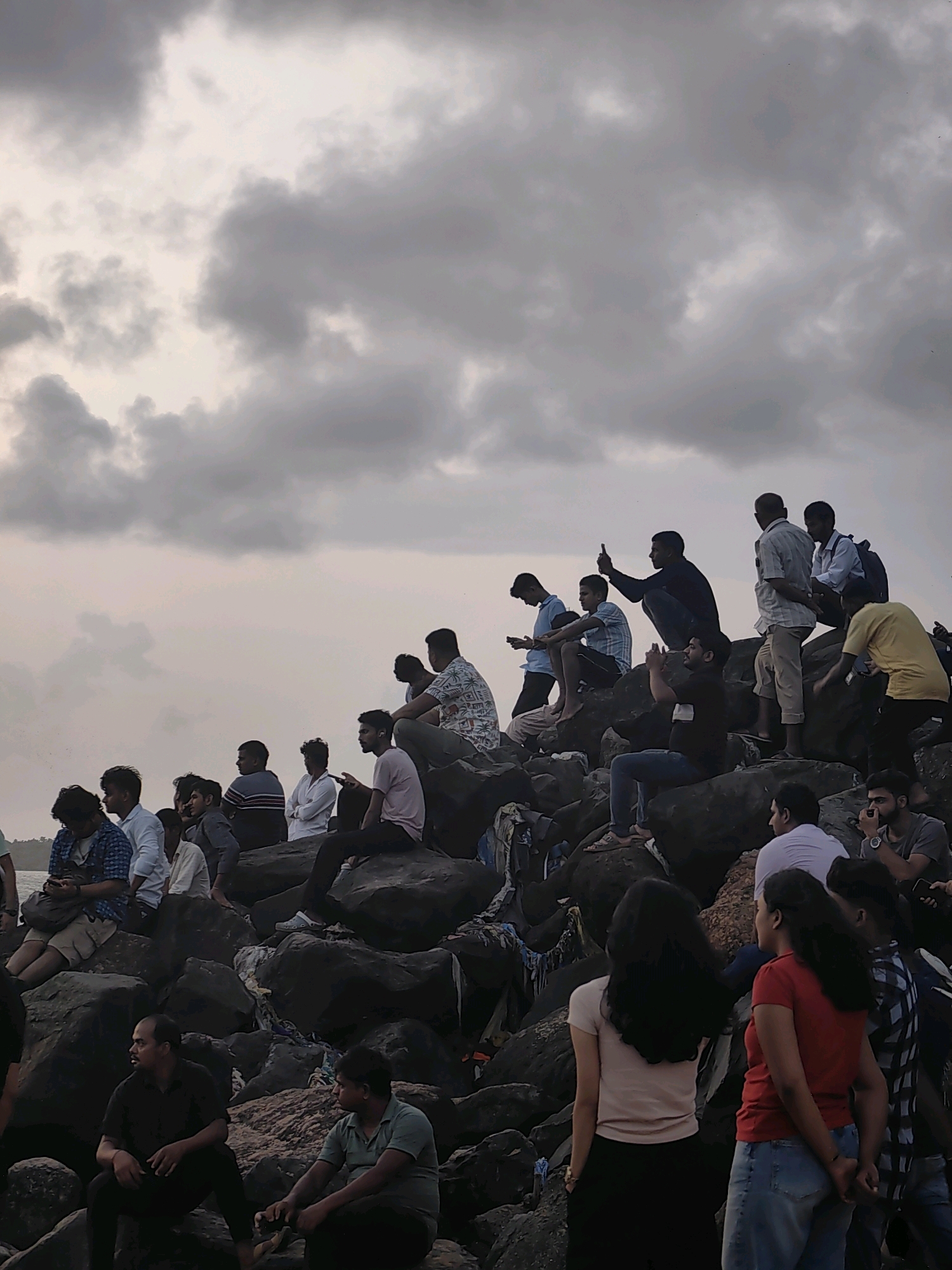 The image captures a group of people gathered on a rocky shoreline, likely in Mumbai, India, given the commonality of such locations for leisure. The sky above is overcast with dramatic, dark clouds, suggesting an approaching storm or late afternoon light. The people are diverse in age and gender, with many sitting and standing on the uneven rocks, some appearing to be in conversation, others looking out towards the water or engaged with their phones. Clothing ranges from casual t-shirts and shirts to more formal attire. The rocky terrain is varied, with some areas appearing worn smooth by the elements, while others are jagged. Scattered debris, possibly plastic bags and cloth, is visible between the rocks in the foreground, hinting at human presence and potentially a public recreational area. The overall atmosphere is one of a casual gathering in a natural, albeit somewhat rugged, urban coastal setting.