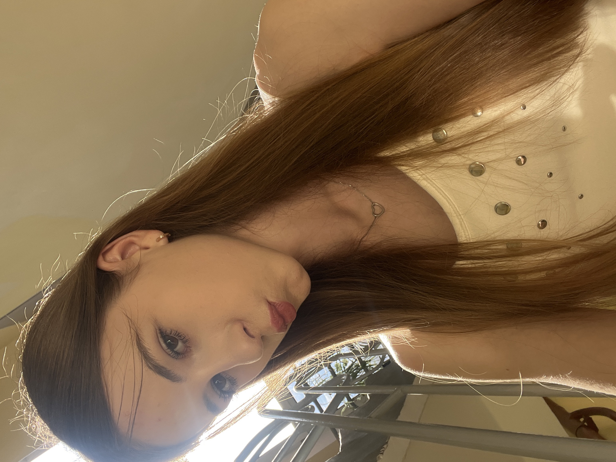A young woman with long, brown hair is captured in a close-up, upside-down selfie. She is wearing a white top adorned with silver circular embellishments. A delicate silver necklace with a heart-shaped pendant rests on her collarbone. Her left ear is visible, featuring a small hoop earring. The lighting suggests it is daytime, with sunlight casting a warm glow and creating highlights on her hair and skin. The background is softly blurred, showing hints of greenery and a metal railing, possibly indicating an outdoor setting like a balcony or garden in Dnipro, Ukraine. The woman's expression is relaxed and contemplative.