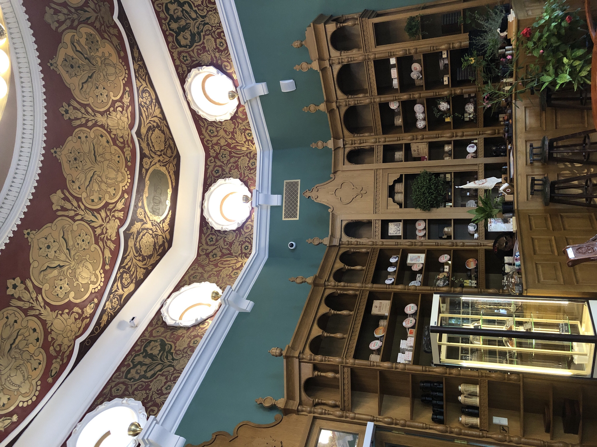 The image shows the interior of a store with ornate decorations and wooden shelving. The ceiling features intricate gold and red patterns with round light fixtures. The walls are painted a teal green. The store has extensive wooden shelving filled with various items, including what appear to be tea canisters, plates, boxes, and some potted plants. A glass display case is visible, illuminated from within, showcasing more items. A wooden counter is present, with stools in front of it. The overall impression is one of a well-decorated establishment, possibly a tea shop or a store selling traditional goods. The mention of Moscow, Russia, is not evident from the visual cues in the image itself.