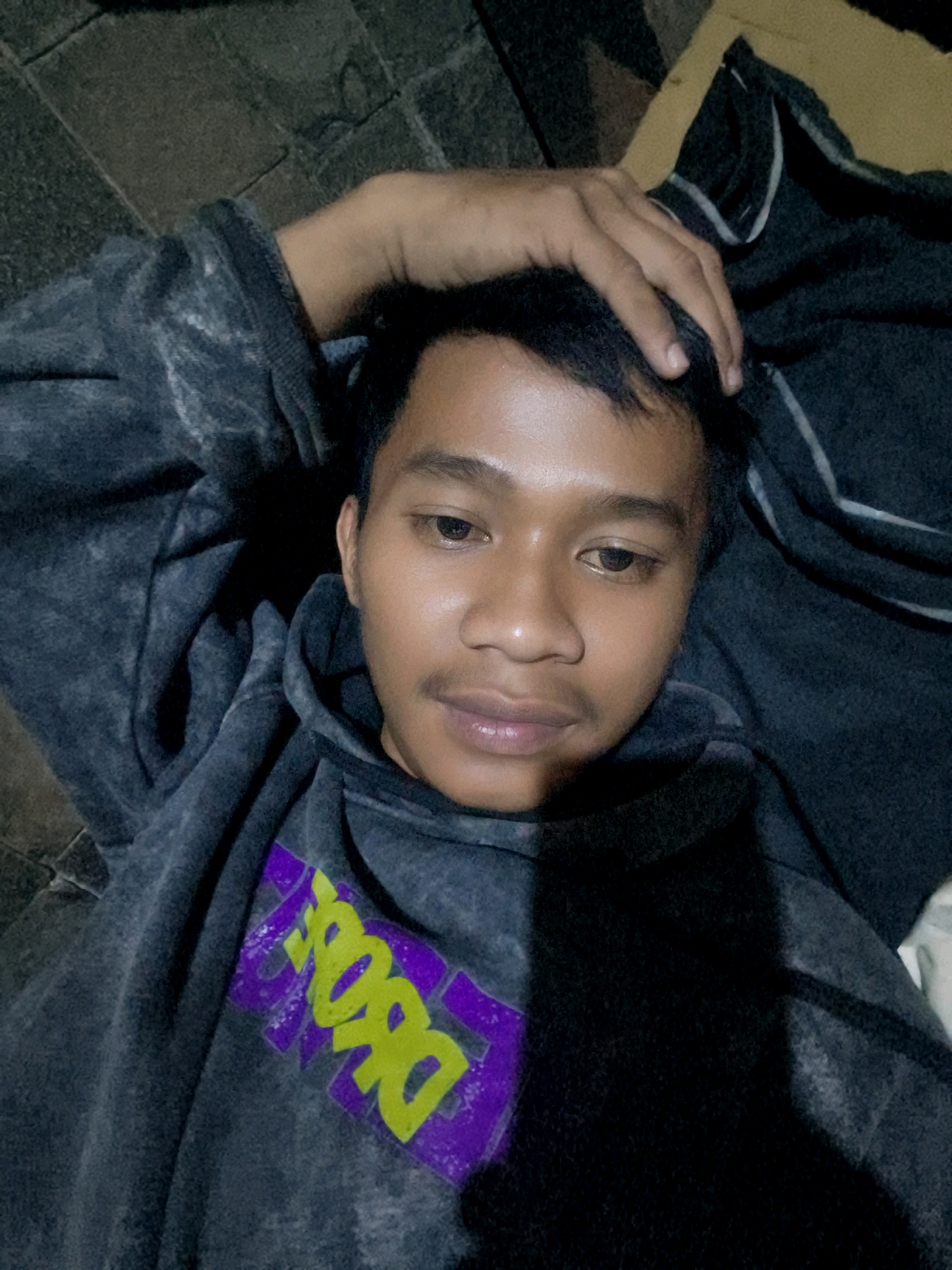 The image shows a close-up, overhead shot of a young man lying down, possibly on a tiled floor, with a blanket or garment draped over him. He is wearing a dark-colored hoodie with a graphic design on the chest featuring purple and neon yellow lettering that appears to say "DOPE". His right hand is placed on his head, touching his hair. The lighting is dim, suggesting it might be nighttime or an indoor setting with minimal light. The overall atmosphere is casual and relaxed. The image does not provide any specific visual cues to pinpoint its location within the city of Bekasi, Indonesia.