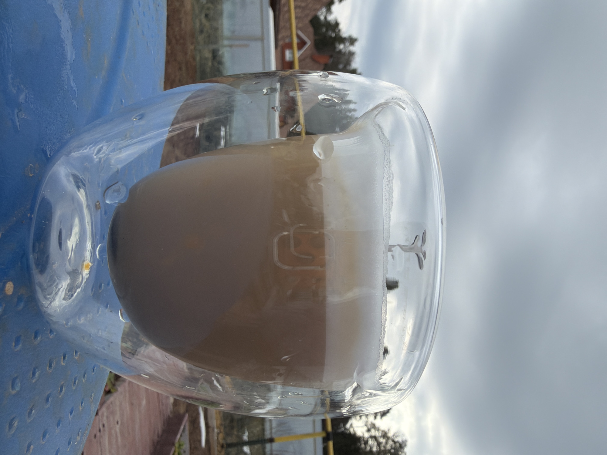 The image shows a glass mug with a layered coffee drink, possibly a latte or cappuccino. The mug is made of clear glass with a double-walled design. The drink has a dark brown liquid at the bottom, topped with a layer of lighter, frothy milk or foam. A small metal stirrer or spoon is inside the mug.
The mug is placed on a blue, possibly plastic, surface. The background is blurred and shows vegetation, parts of a building, and an overcast sky.
There is no information available from the image to determine the location is Chaikovsky, Russia.
