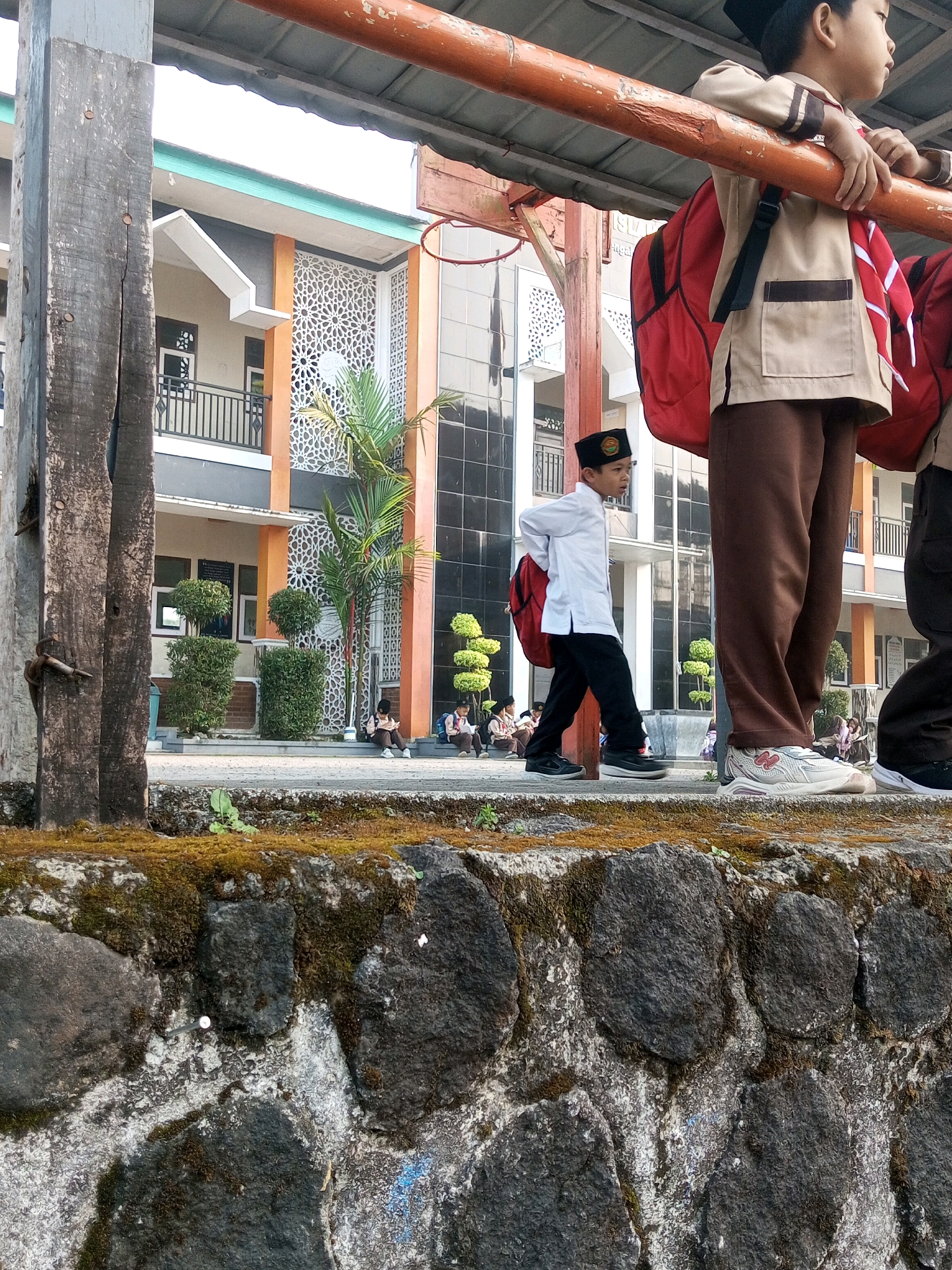 The image depicts an outdoor scene in front of a school building in Kalibeber, Indonesia. The foreground is dominated by a textured stone wall covered in moss and some dry grass. Behind the wall, a paved walkway is visible, and further back, a building with modern architecture featuring orange and white decorative panels and large windows is present. Several students in uniform are visible. In the mid-ground, a young boy in a white shirt, brown pants, and a black cap is walking with a red backpack. To the right of him, the legs and lower torso of another student in brown pants and white sneakers are visible. Further in the background, several other students are seated or walking. The overall environment suggests a school setting during the daytime.