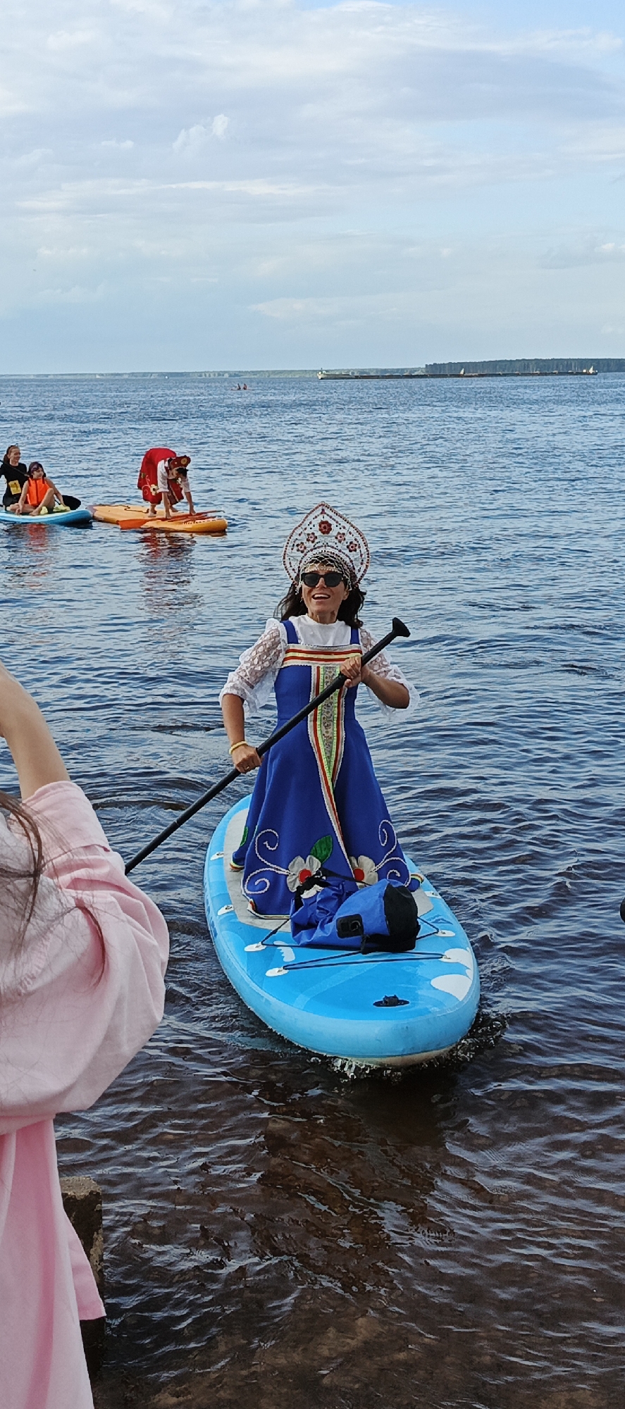 A woman in traditional Russian attire, including a kokoshnik headdress and a blue dress with floral embroidery, stands on a blue paddleboard in a body of water, holding a paddle. She is wearing sunglasses and smiling. She is on the Kama River in Chaikovsky, Russia. In the background, other people are on paddleboards, and a barge is visible on the horizon. The sky is cloudy.