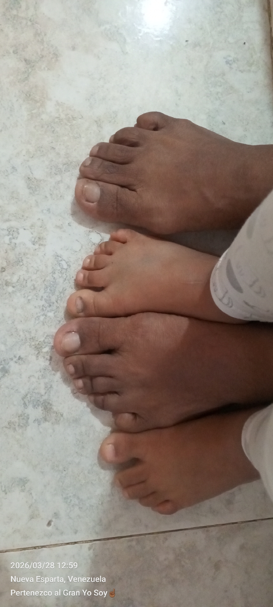 The image shows two pairs of bare human feet resting on a tiled floor. One pair appears to be larger and darker in skin tone than the other. The feet are partially overlapping. A portion of fabric is visible near the feet. The image's timestamp indicates it was taken on March 28, 2026, at 12:59 in Nueva Esparta, Venezuela. Altagracia is not explicitly mentioned in the text.