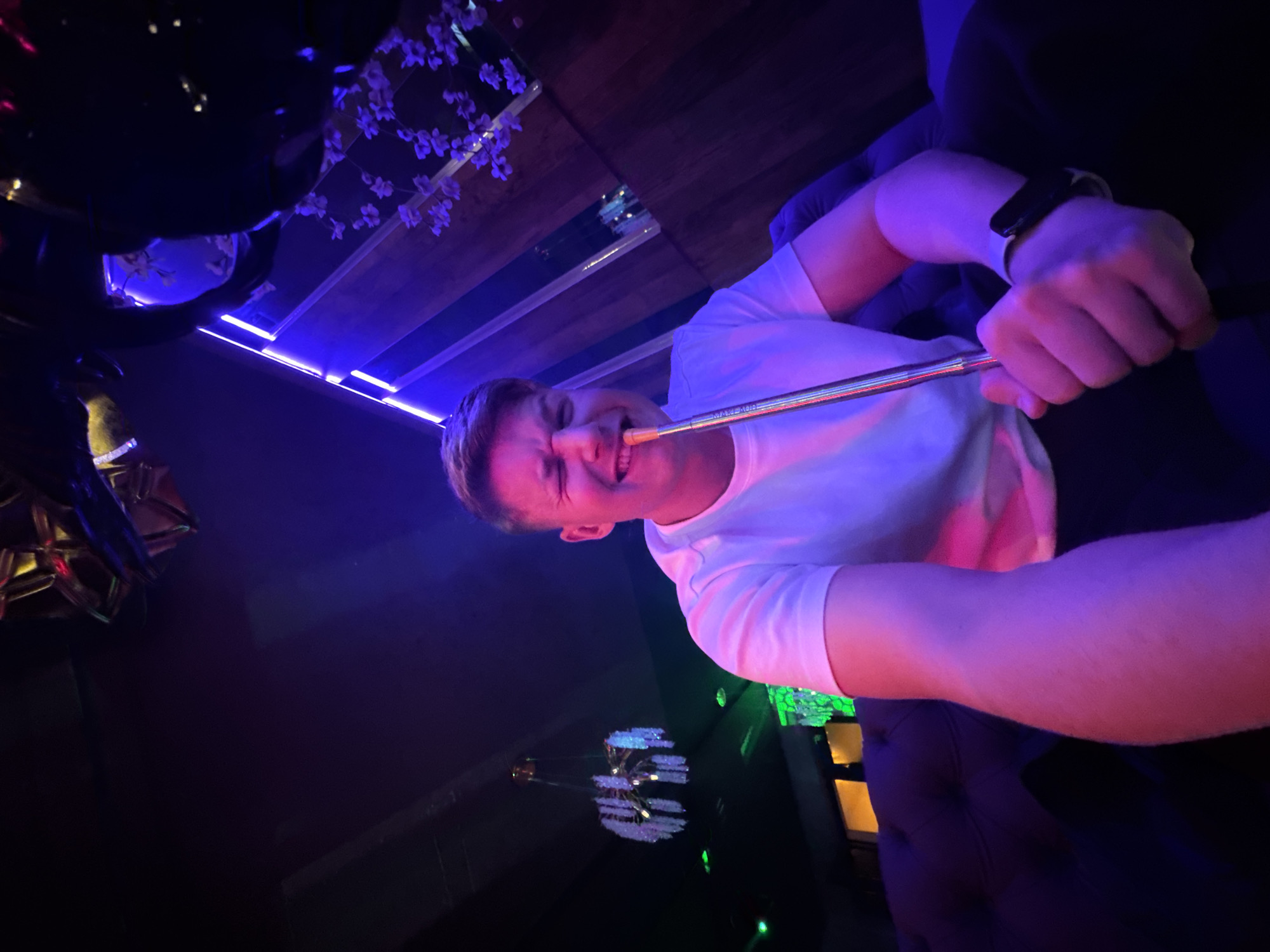 A fair-skinned person is smoking a hookah pipe in a dark room. The person has short, light-colored hair and is wearing a white t-shirt and a smartwatch on their left wrist. The hookah pipe is silver with a dark mouthpiece. The room has a modern, dark decor with blue and purple lighting, a mirrored ceiling with decorative white flowers, and a chandelier. It's indoors, potentially in a bar or lounge. The location is Burgas, Bulgaria.