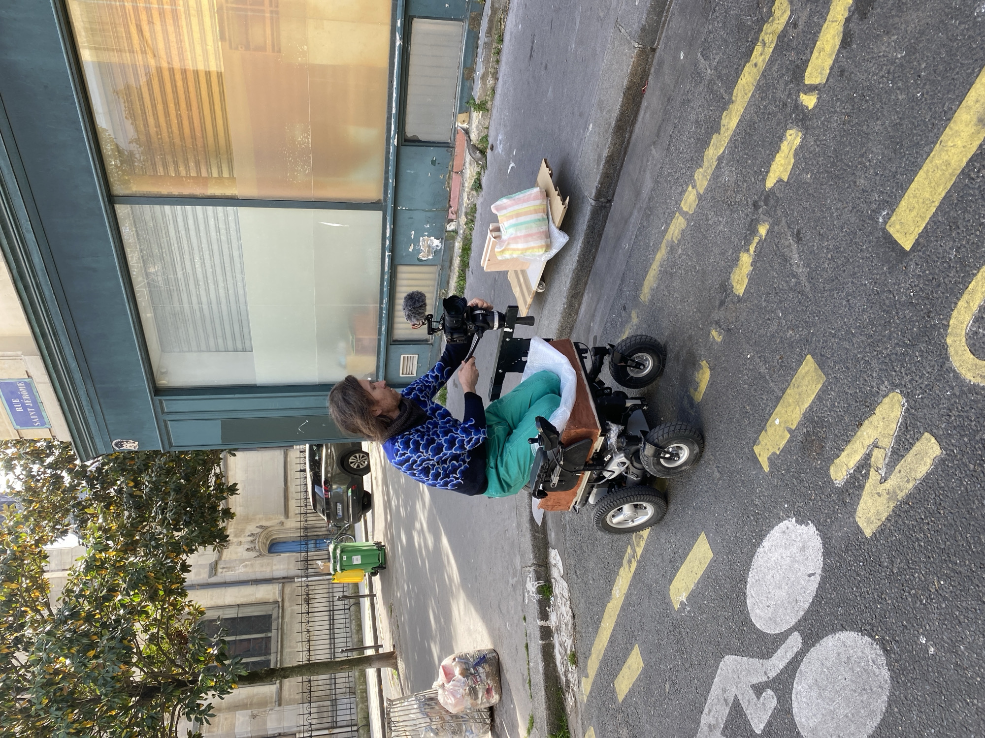 An urban street scene unfolds on a bright, sunny day in Paris, France. The central subject is a person with long brown hair, wearing a blue and white patterned long-sleeved top and green trousers, operating a professional camera. They are seated on a low-slung, customized four-wheeled motorized cart, possibly an adapted mobility scooter, which features a wooden seat. The camera, equipped with a large lens and a furry microphone, is mounted on a small rig in front of them, suggesting they are engaged in filming or recording.
The cart is positioned on the edge of an asphalt road marked with yellow dashed and solid lines, as well as a white painted symbol indicating a bicycle path. To the right of the person, a cardboard box containing folded textiles sits on the sidewalk near the curb. Behind the person, a building with large, somewhat aged windows reflecting the bright sky forms the immediate backdrop. Further back on the left, a mature tree with dark green leaves partially obscures a classic stone building.
On the opposite side of the street, more traditional Parisian architecture is visible, including a building with an arched window. A black car is parked along the curb, and green and yellow recycling bins stand near the building. Closer to the foreground on the sidewalk, a clear plastic bag filled with waste is placed beside a metal grate. A blue street sign, partially visible, reads "RUE SAINT JEROME." The overall impression is one of an active street environment, likely captured during a film shoot or similar creative endeavor.