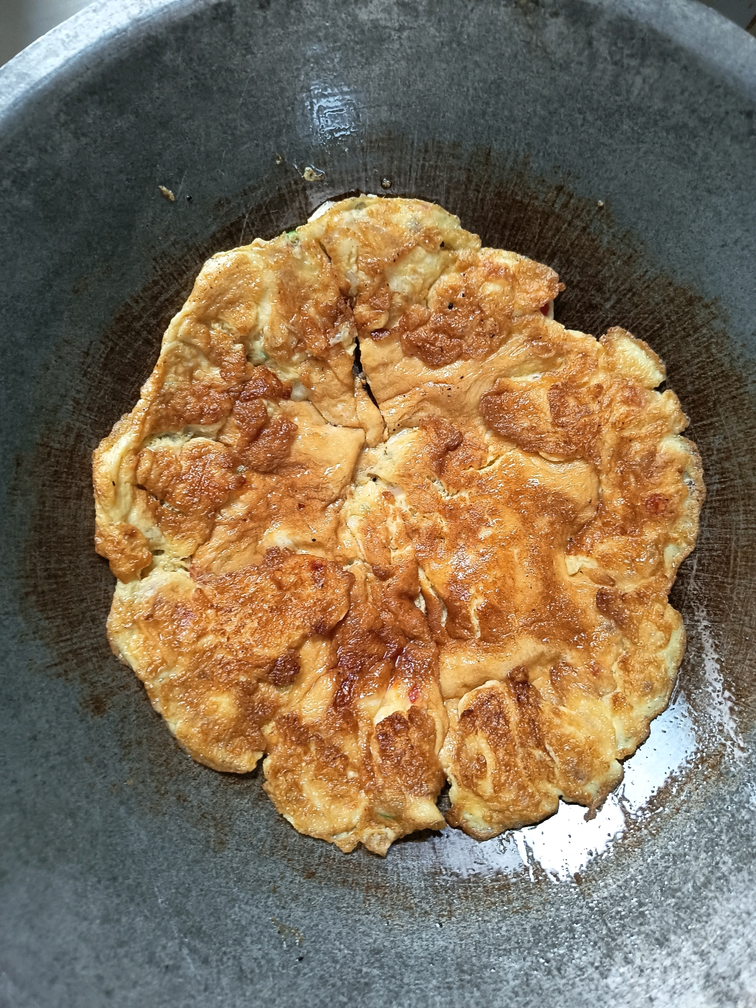 A close-up overhead shot shows a golden-brown, slightly uneven omelet cooking in a dark, round pan. The omelet has a textured surface with some darker browned areas, indicating it is well-cooked. The pan appears to be made of metal and has a slightly worn, possibly seasoned, surface. There are some shiny spots, likely from oil, on the pan around the omelet. The background is filled with the dark gray of the pan. This image is located in Aluekumba, Indonesia.
