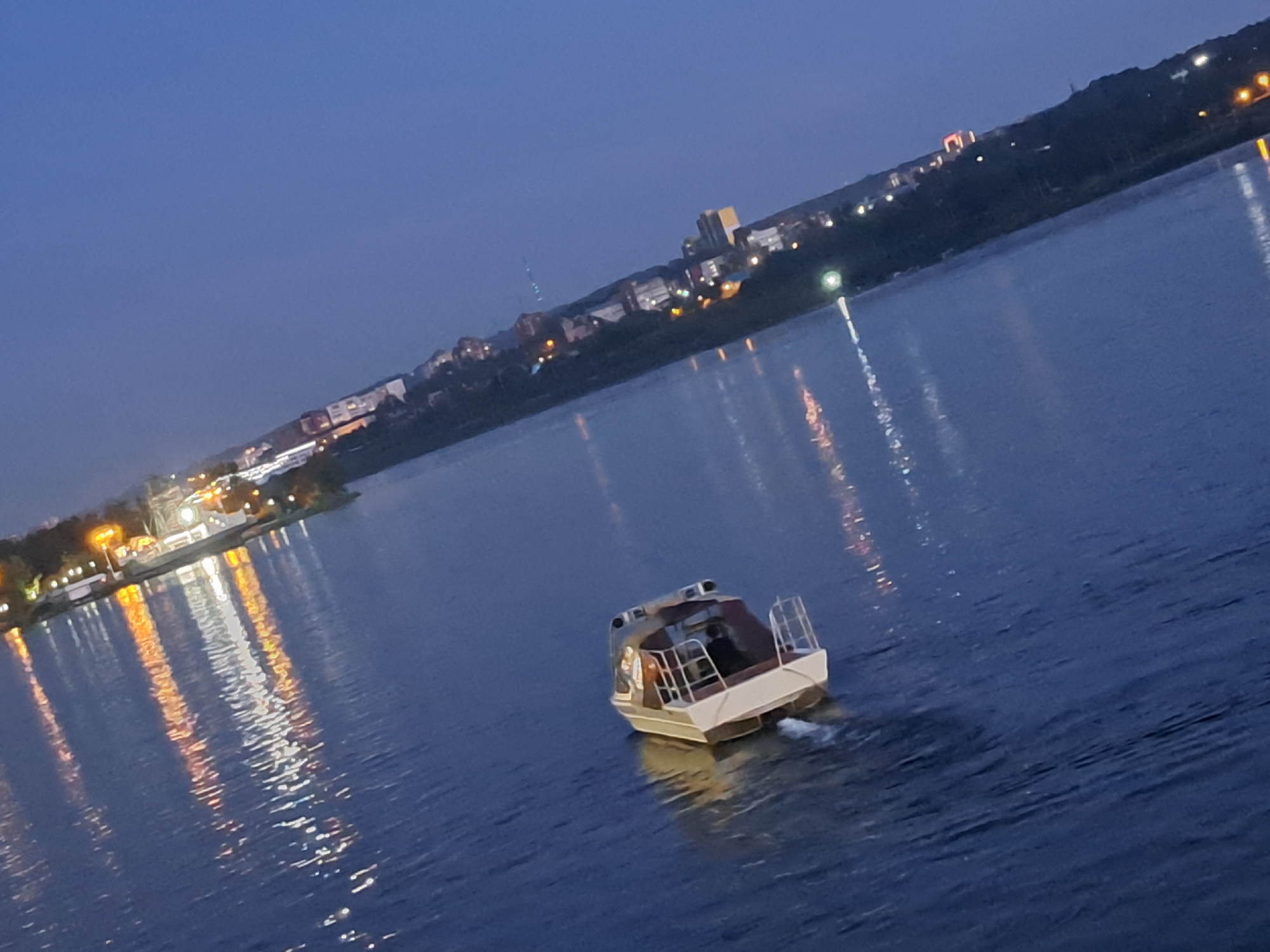 A motorboat travels on a dark blue body of water, leaving a white wake. The boat is light-colored with a darker cabin and railing, and appears to have at least two people inside. The background shows a city skyline at dusk, with numerous lights reflecting on the water. The city appears to be Irkutsk, Russia, with lights visible along the shoreline and buildings on a slope overlooking the water. The overall atmosphere is calm and serene, with the lights creating a vibrant contrast against the twilight sky.