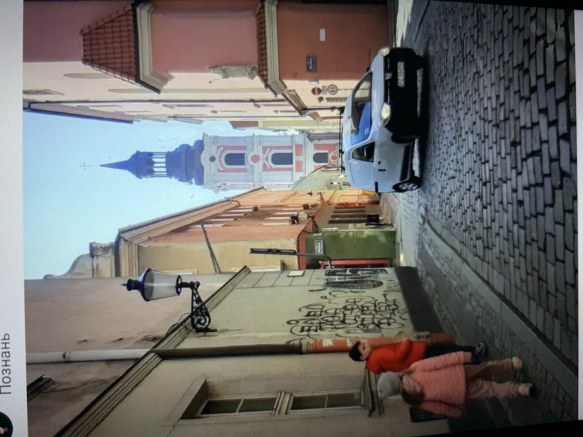 This image depicts a narrow, cobblestone street scene in what appears to be a European city. The architecture suggests an older urban environment with buildings of varying heights and colors lining the street. A prominent feature in the background is a tall church tower with a distinctive spire, characterized by white and pink detailing. In the foreground, a white van is parked on the street. Two children are visible on the left side of the street, one in a red jacket and the other in a pink outfit, facing away from the camera. The walls of the buildings are marked with graffiti. The sky is clear and blue, indicating fair weather. The image quality is somewhat reduced, possibly due to being a photograph of a screen. The text overlay at the top of the image is in Cyrillic script.