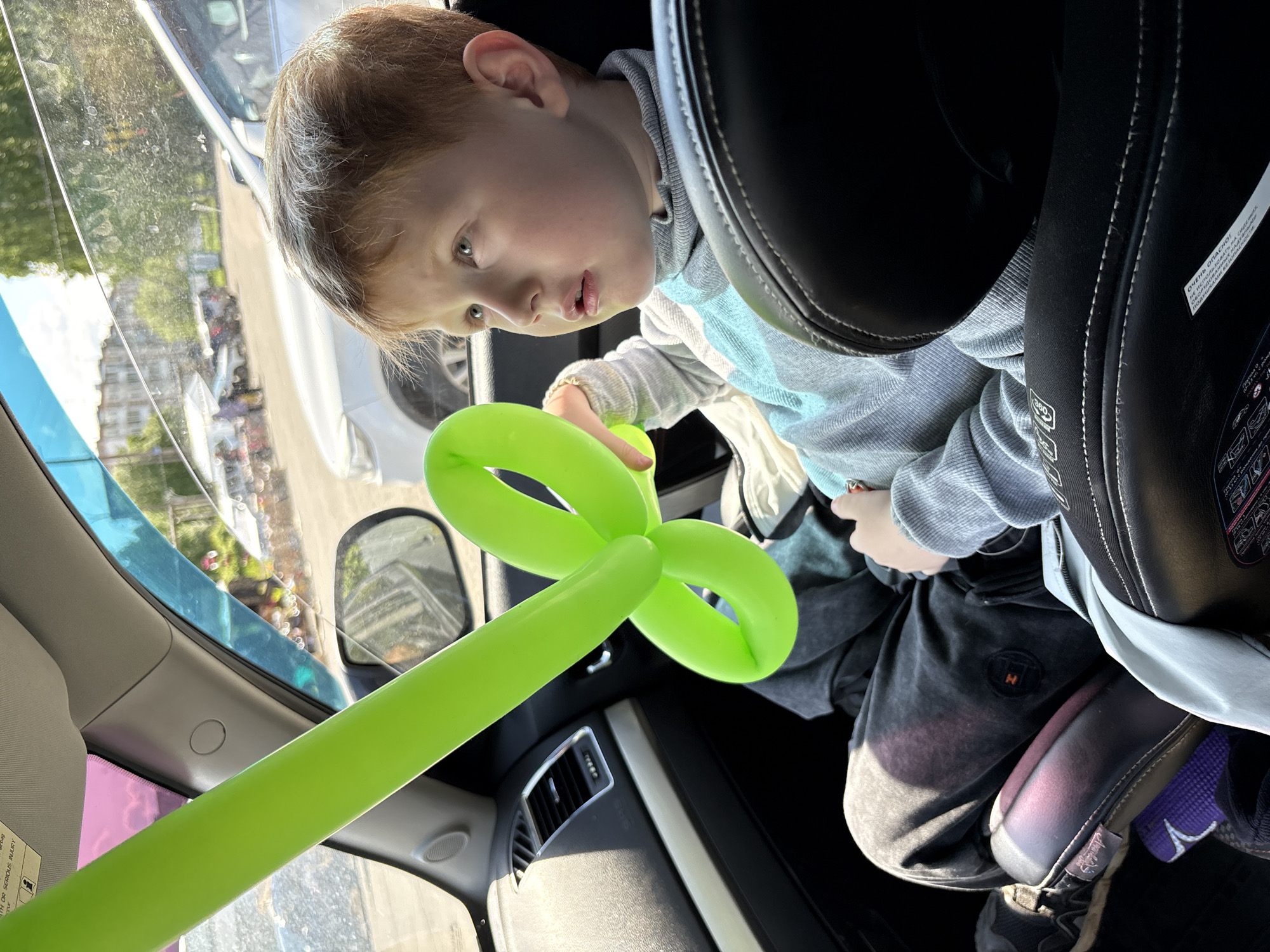 A young child is seated in a car seat, inside a vehicle, holding a green balloon animal in the shape of a flower or clover. The child is wearing a grey sweatshirt and dark pants. The vehicle appears to be parked, with a view of a street and buildings visible through the window. There's no evidence to suggest this image was taken in the city of Италмас, Russia.