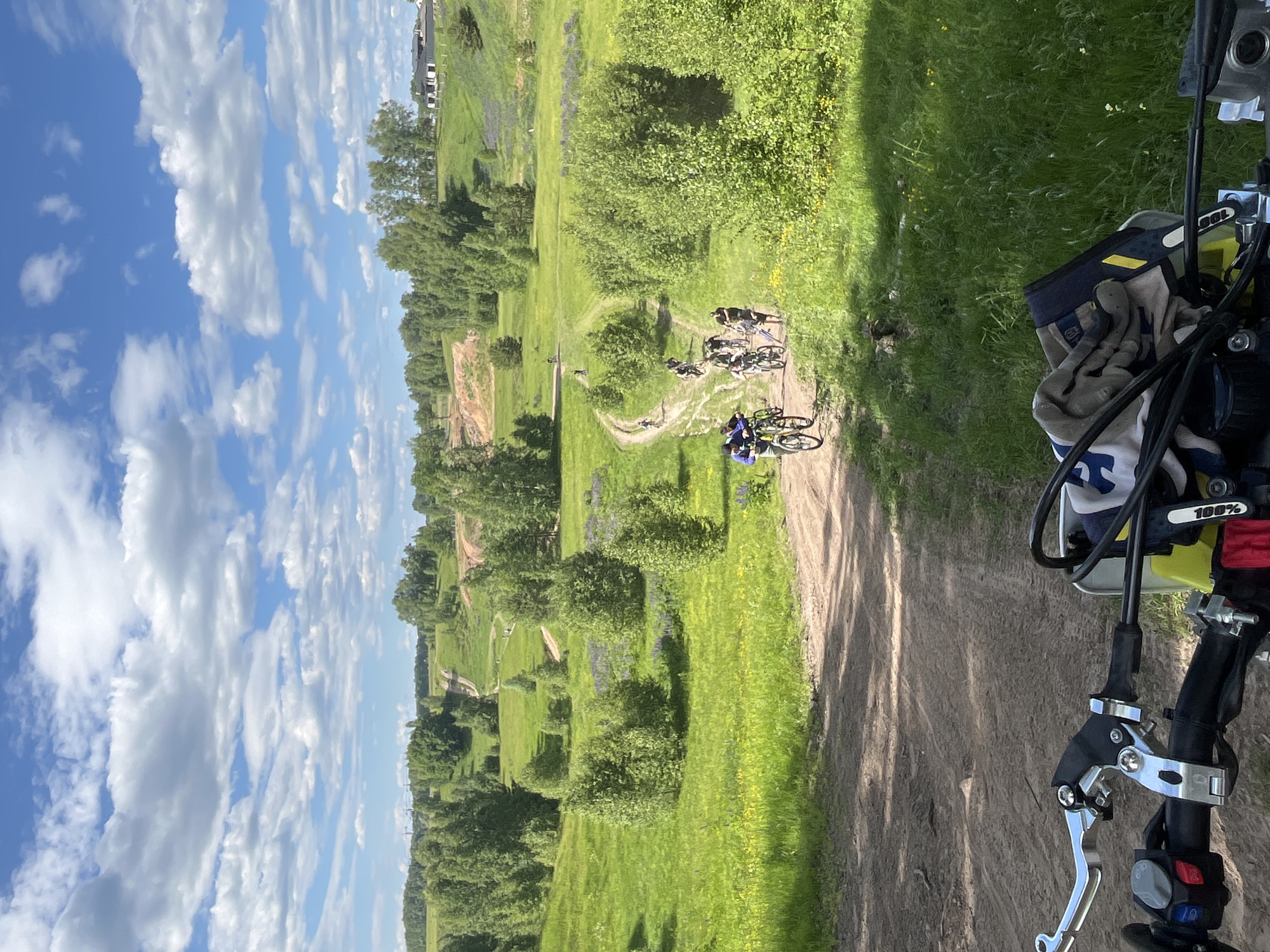 This outdoor daytime image, captured in the vicinity of Novochnuvashsky, Russia, depicts a group of cyclists navigating a series of unpaved dirt paths through a verdant, rolling landscape under a bright blue sky with scattered white cumulus clouds.
In the distant background, gentle hills are covered with dense deciduous forests, with a few white residential-style buildings topped with dark roofs visible on a distant crest. Far left, faint industrial structures or power lines are discernible.
The midground is characterized by expansive green fields interspersed with patches of purple and yellow wildflowers. Numerous small, bushy deciduous trees are scattered throughout the fields and along the winding dirt paths. A group of approximately ten individuals, primarily on mountain bikes, are seen traversing these paths, generally moving away from the viewer. One cyclist in the foreground group wears a blue jacket and stands next to a yellow mountain bike. Other cyclists are visible further along the paths, navigating turns and inclines.
The image is taken from the first-person perspective of a person on a motorcycle or dirt bike, with the vehicle's handlebars prominently featured across the bottom of the frame. Visible components include a silver-colored clutch lever assembly on the left, black rubber grips, and left-hand controls with red, blue, and gray buttons/switches. A pair of off-white and blue patterned gloves or a cloth, bearing a "100%" logo, is draped over the right side of the handlebars. The immediate foreground is a dirt track, leading into the broader path network.