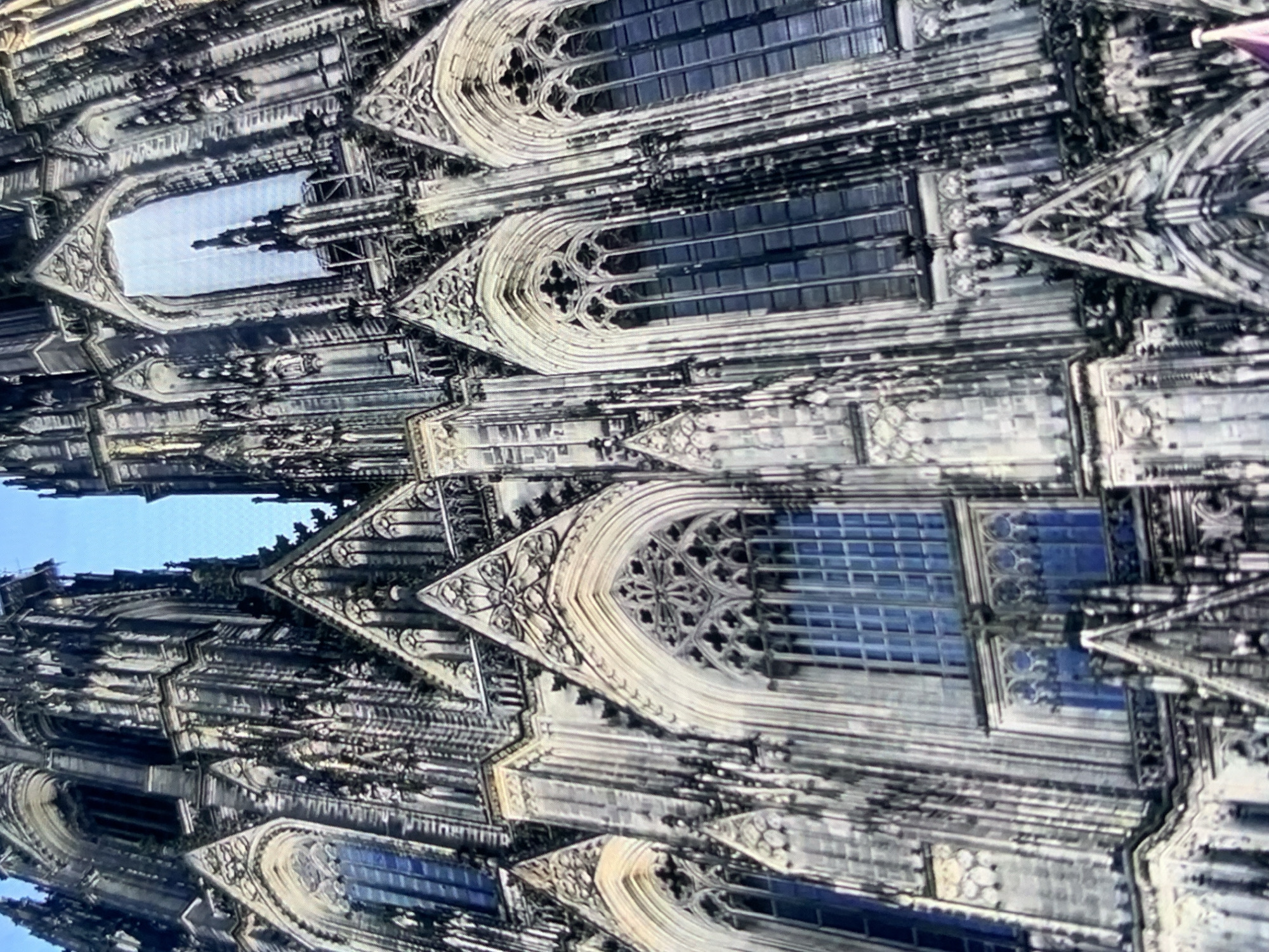 The image shows a close-up, low-angle view of the facade of a large Gothic cathedral, characterized by intricate stonework, pointed arches, and tall spires. The cathedral appears to be constructed of dark stone, with many ornate details and sculptures. Several large stained-glass windows are visible, reflecting a blue sky. The sky above is clear and bright blue. The scene is a daytime shot, with natural light illuminating the cathedral's architecture. This image does not depict Stavropol, Russia.
