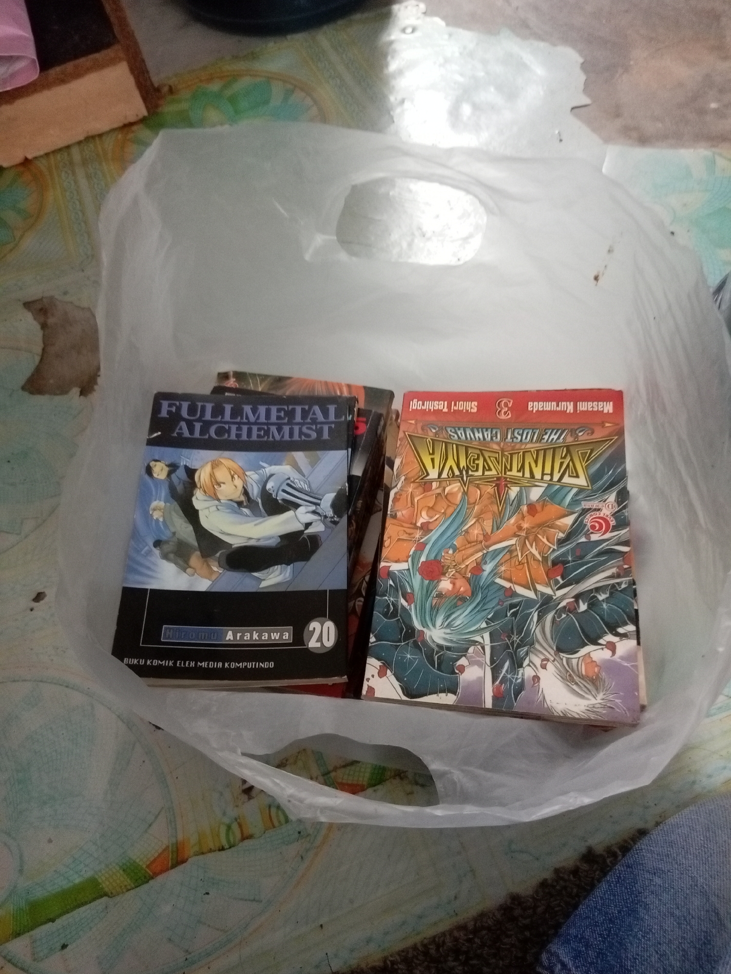 Two manga books are visible within a white plastic bag. The book on the left is "Fullmetal Alchemist" Volume 20 by Hiromu Arakawa. The book on the right is "Saint Seiya The Lost Canvas" Volume 3 by Masami Kurumada and Shiori Teshirogi. The bag is placed on a patterned surface, possibly a mat or floor, with some scattered debris around it. The background suggests an indoor setting, possibly a home environment. The image was taken on March 22, 2024, at 1:00 PM. The location is in Batam, Indonesia.