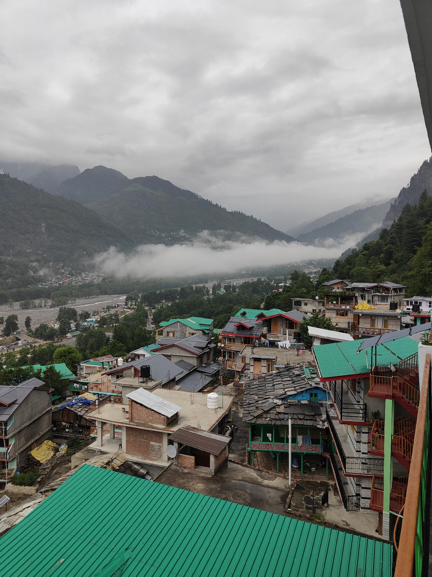 This is a high-angle, full-frame, eye-level shot of a mountainous landscape with a village nestled in the valley, likely Vashisht, India. The sky is overcast with thick, gray clouds. In the distance, verdant mountains rise steeply, their peaks partially obscured by low-lying mist that drifts through the valley. A riverbed, appearing wide and stony, snakes through the valley floor, with a town spread out on its banks, characterized by a collection of buildings with various colored roofs. Closer to the foreground, the scene transitions to a densely packed residential area. Buildings are multi-story structures with a mix of architectural styles, many featuring prominent green metal roofs. Some buildings exhibit balconies with railings, and windows are visible. Various objects are scattered on rooftops and around the buildings, including water tanks, satellite dishes, tarpaulins, and laundry. The immediate foreground is dominated by a large, green corrugated metal roof. The overall impression is one of a somewhat rugged, densely populated settlement situated within a dramatic natural setting. There are no discernible people or specific actions taking place in the image.
