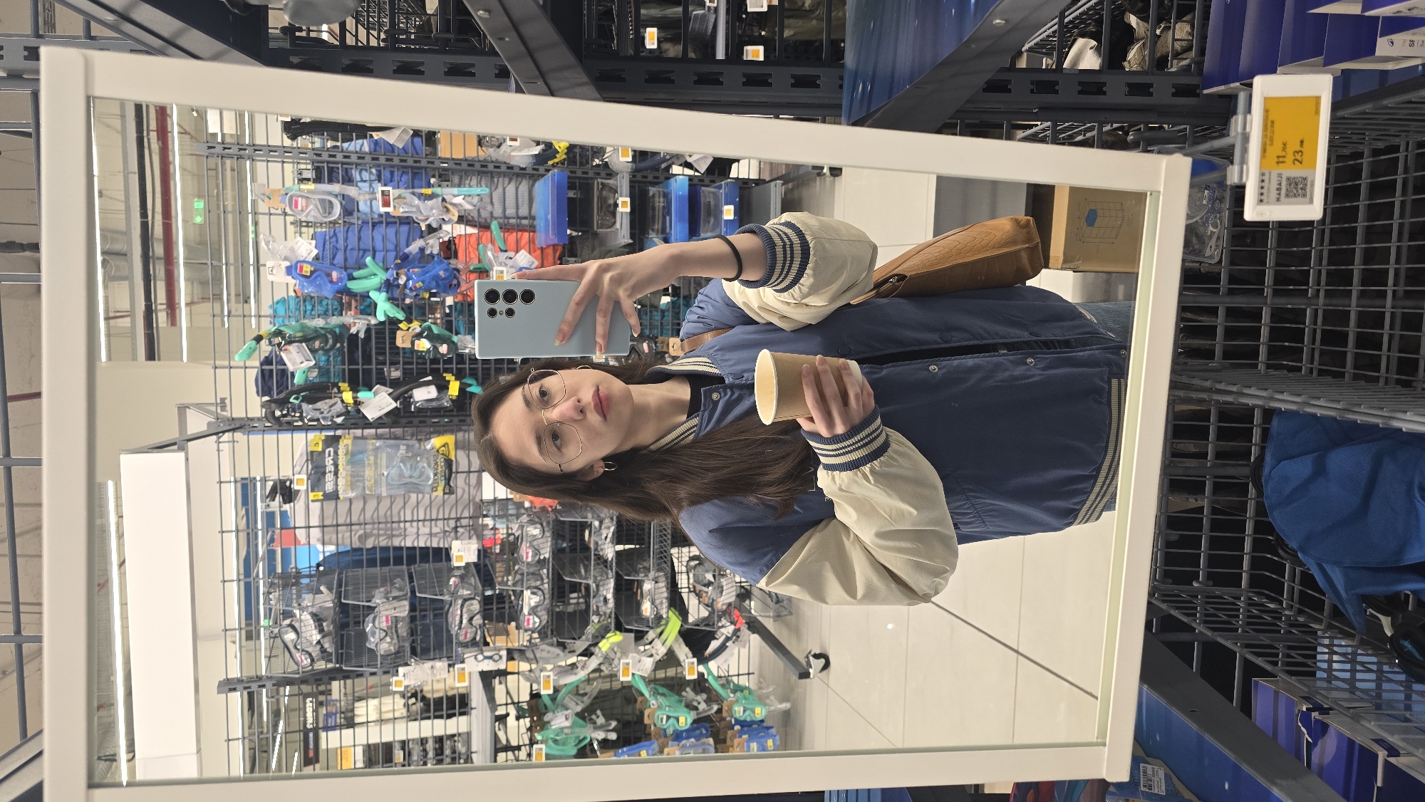 A mirrored selfie shows a young woman standing in a Decathlon store, likely in Stara Zagora, Bulgaria. The woman is holding a light blue smartphone in her left hand, angled to capture her reflection. She holds a small, brown paper cup in her right hand. The woman wears glasses and a blue varsity jacket with beige sleeves. The store's shelving units, stocked with various sporting goods, form the background. A price tag is visible in the upper right corner of the mirror. The floor appears to be white tile.