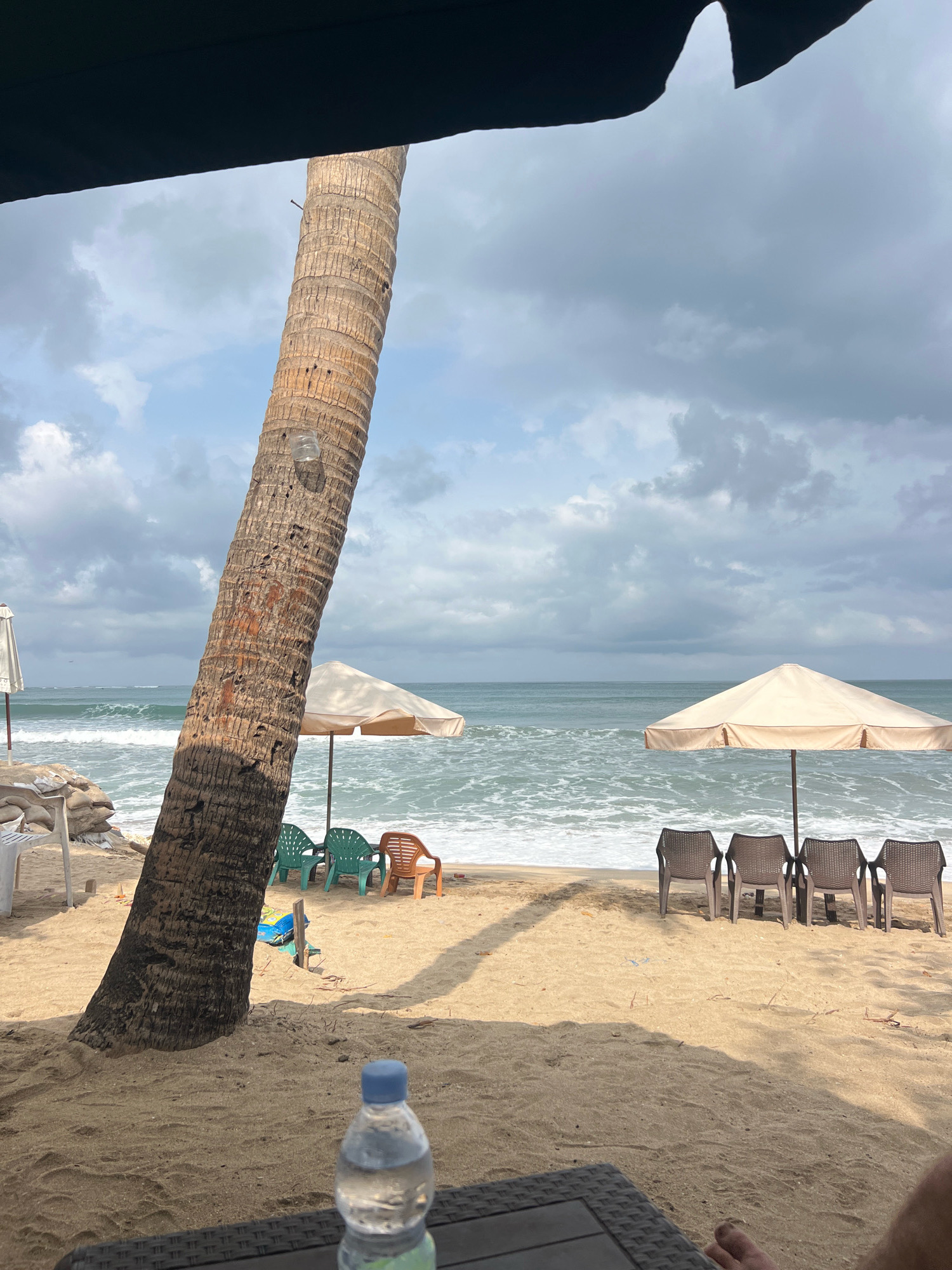 The image depicts a sandy beach in Kuta, Indonesia, under a cloudy sky. In the foreground, a partially visible person's feet and a bottle of water are on a dark surface, possibly a table. A large palm tree trunk dominates the left side of the frame, leaning towards the right. Behind the tree, two beach umbrellas are set up on the sand. The one on the left is partially obscured by the palm tree and appears white. The one on the right is a light beige color and stands open.
In front of the beige umbrella, four dark brown plastic chairs are arranged. In front of the white umbrella, three green plastic chairs and one orange plastic chair are visible. The ocean is in the background, with moderate waves breaking on the shore. The water appears greenish-blue. On the far left of the image, there are some sandbags and a white structure, possibly part of a building or another umbrella. The overall scene suggests a relaxed atmosphere on a somewhat overcast day at the beach.