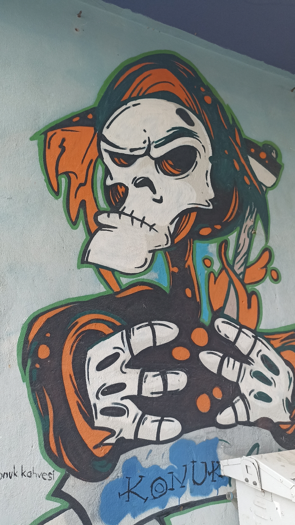 A vibrant graffiti art piece depicting a stylized skull figure is prominently displayed on a light blue wall in Yenişehir, Turkey. The central subject is a white skull with large, dark eye sockets and a stitched jawline, conveying an intense expression. It appears to wear a dark green hood or head covering, adorned with orange patterns, and its body is rendered in black with swirling orange accents and dots. Two large, white skeletal hands with distinct finger bones are positioned in the foreground, near the bottom of the figure.
The graffiti employs a bold style with strong black and green outlines, and a distinct color palette of white, black, orange, and dark green against the pale blue wall. The wall itself shows some texture and wear.
At the bottom left of the image, the phrase "konuk kahvesi" (Turkish for "guest coffee" or "visitor's coffee") is written in a neat, green script. Below the graffiti figure's hands, the word "KONUK" (Turkish for "guest" or "visitor") is spray-painted in blue with black outlines, in a more rugged style. A white utility box is partially visible in the bottom right corner of the frame. The top edge of the wall transitions to a darker blue, possibly indicating a corner or ceiling. The lighting suggests daylight conditions, with no harsh shadows.