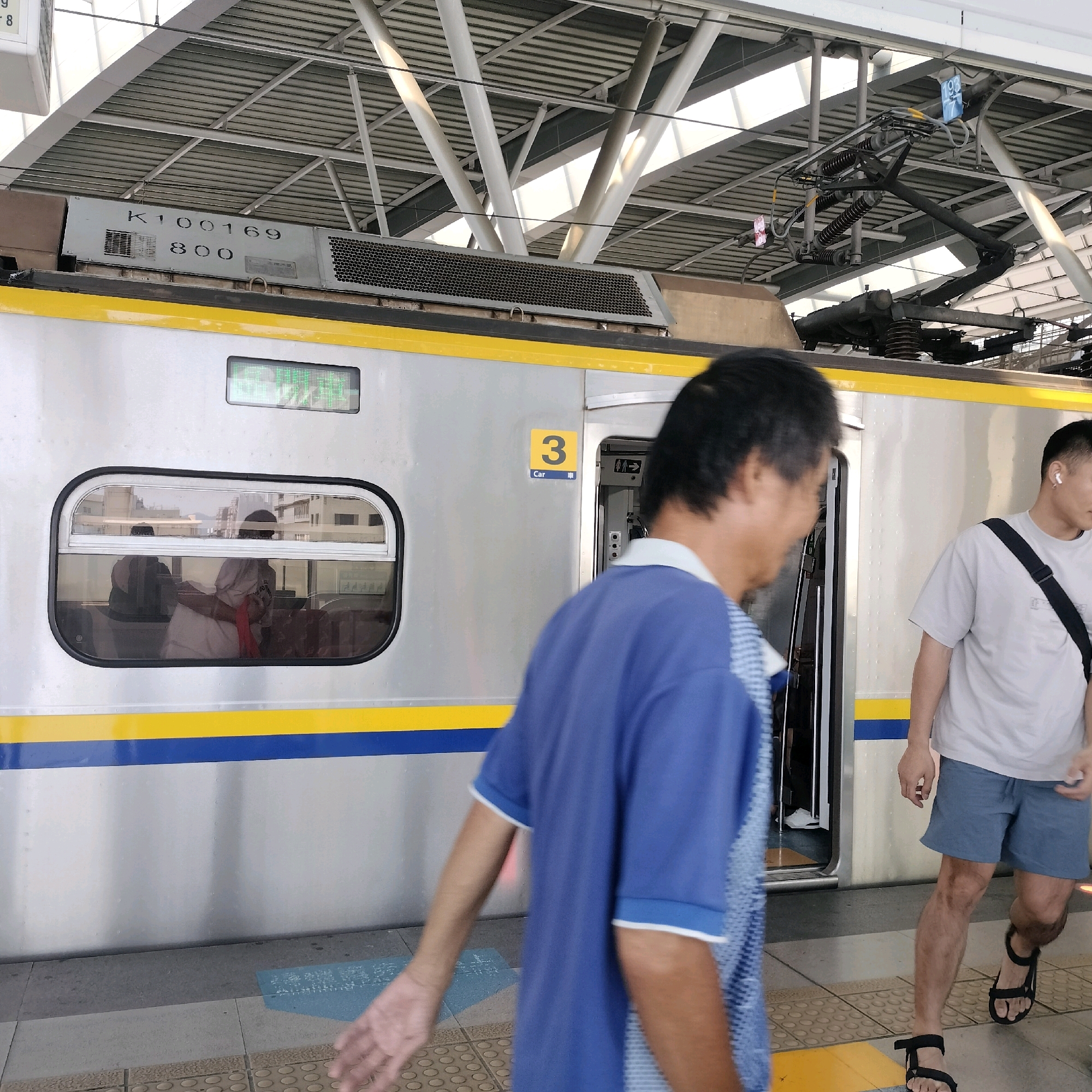 At a Taichung TRA (Taiwan Railways Administration) station, possibly near a station like Taichung Station, a train is stopped at a platform. The train car is silver with a yellow and blue stripe along its side. A digital display above a window shows green text. A sign on the train door indicates "3 Car." People are on the platform, with some boarding or disembarking. The train is modern, featuring an overhead pantograph for electrical power. The platform itself has yellow and gray checkered tiles. The overhead structure of the station consists of a metal roof supported by white pillars. One person, wearing a blue shirt, is partially visible in the foreground, walking away from the camera. Another person in a light-colored t-shirt and blue shorts is standing near the train door, facing towards the right. The lighting suggests it is daytime.