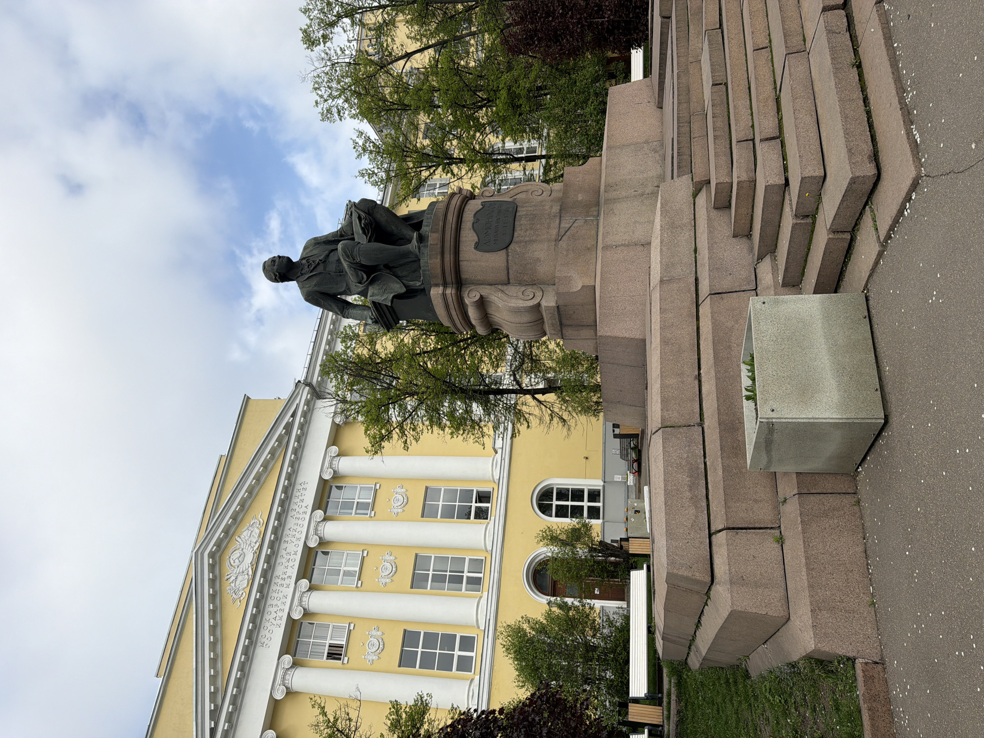 The image shows the statue of Mikhail Lomonosov in Moscow, Russia. The statue is made of dark metal and is set on a stepped base made of light brown stone. Behind the statue is a yellow building with white columns and ornamentation. The sky is partially cloudy. There are trees scattered throughout the scene.