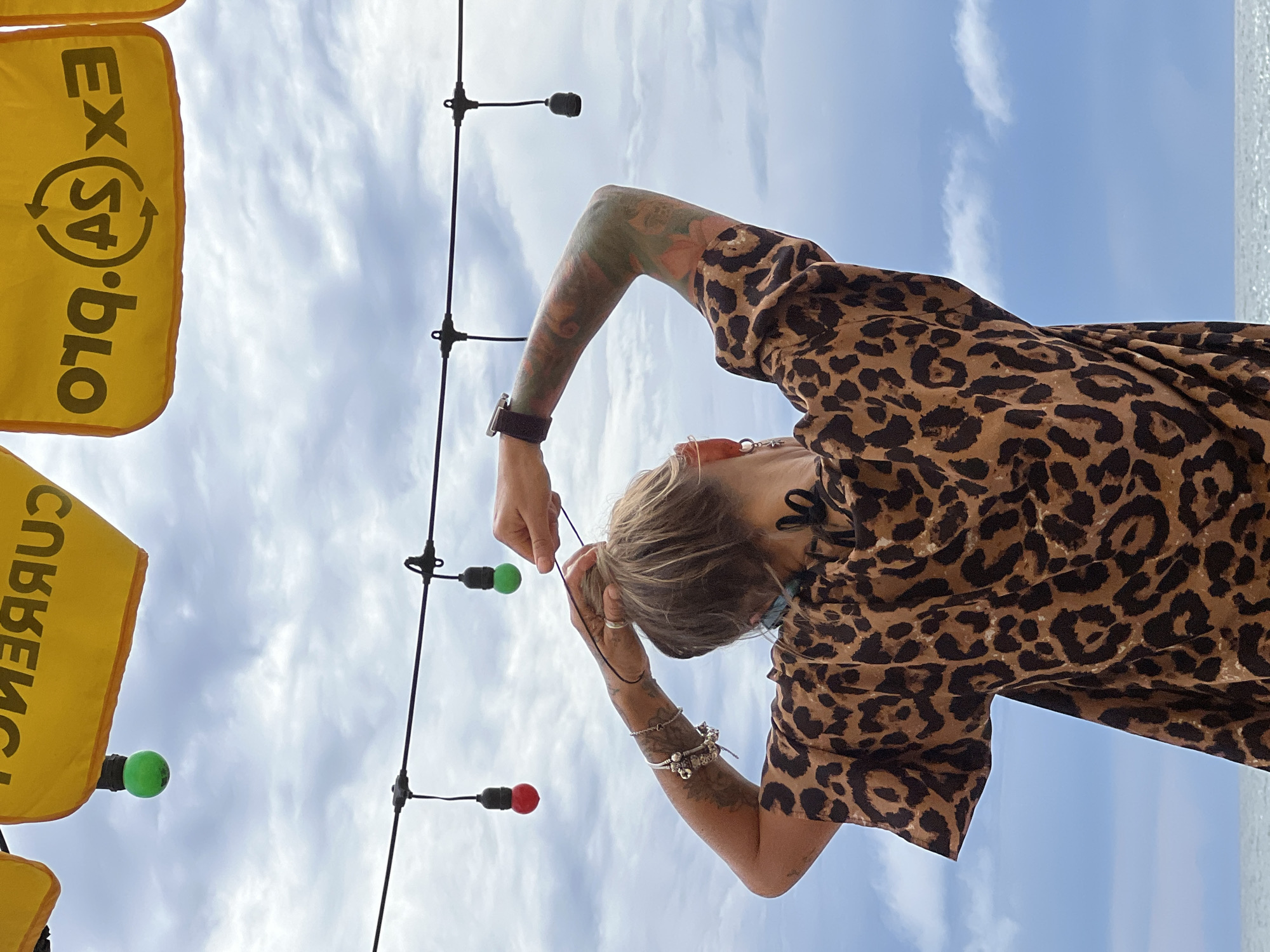 A woman with tattoos on her arms and wearing a leopard-print top is standing with her back to the camera and her arms raised, adjusting her hair. She is outdoors with a cloudy sky and the ocean visible in the background. There are strings of lights hanging above her, some with colored bulbs. Two yellow banners with text are visible at the top of the frame.