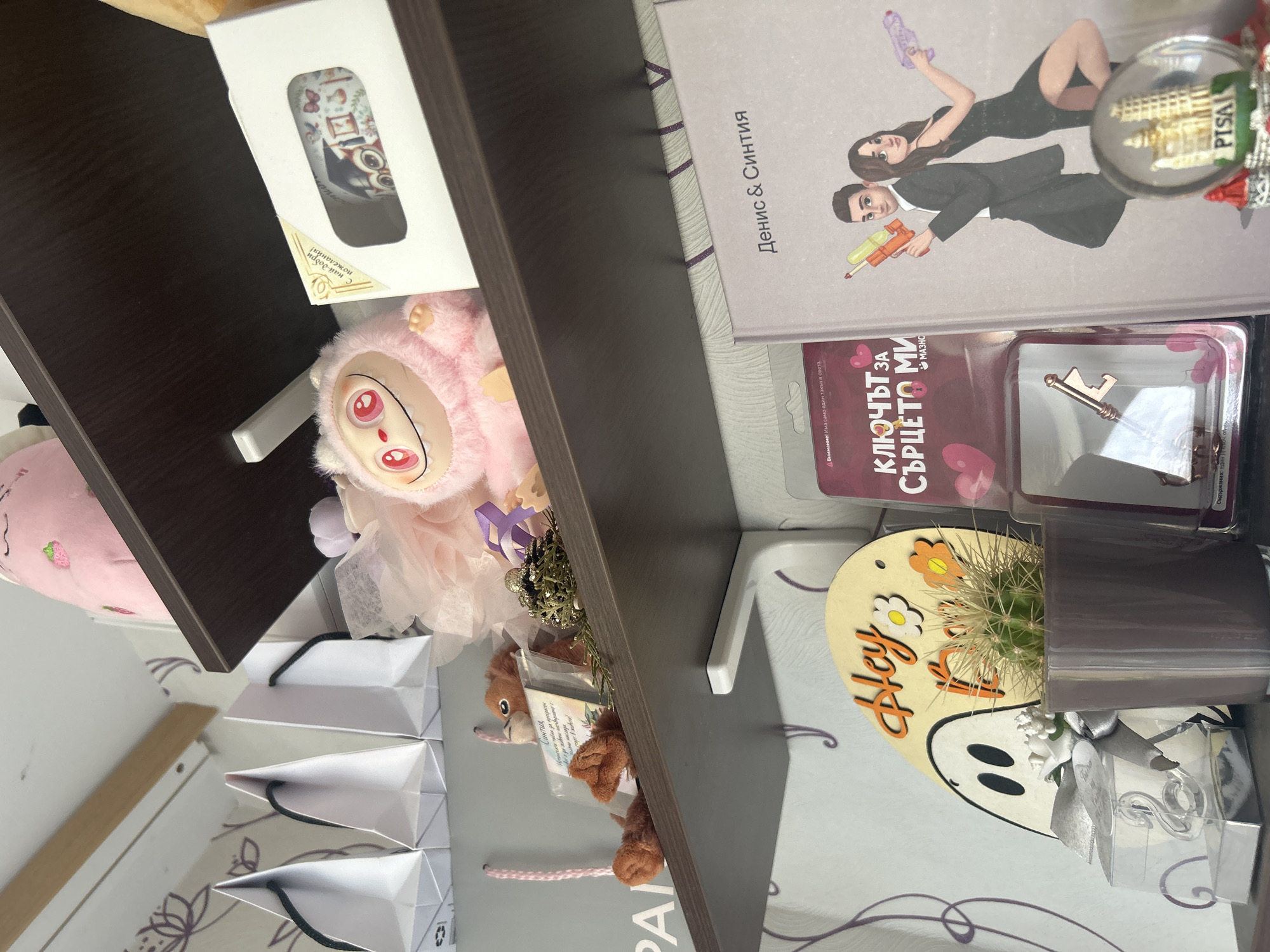 A shelf holds an assortment of items. Starting from the top left, a white box with a decorative design is partially visible, followed by a pink plush toy. Below these items are several small, white shopping bags. Moving towards the right, there's a book titled "Денис & Синтия" (Denis & Cynthia) with a cartoon-like image of a couple holding toy guns. A souvenir snow globe with the Leaning Tower of Pisa is positioned near the book. Underneath the book is a card that reads "КЛЮЧЪТ ЗА СЪРЦЕТО МИ" (Key to My Heart), along with a decorative key. Adjacent to these items is a small cactus in a dark pot, sitting on a decorative plaque that says "Hey". A plush squirrel, and a shopping bag with "Pandora" are also visible. The shelf is dark brown and the background includes a white surface with a lavender pattern.