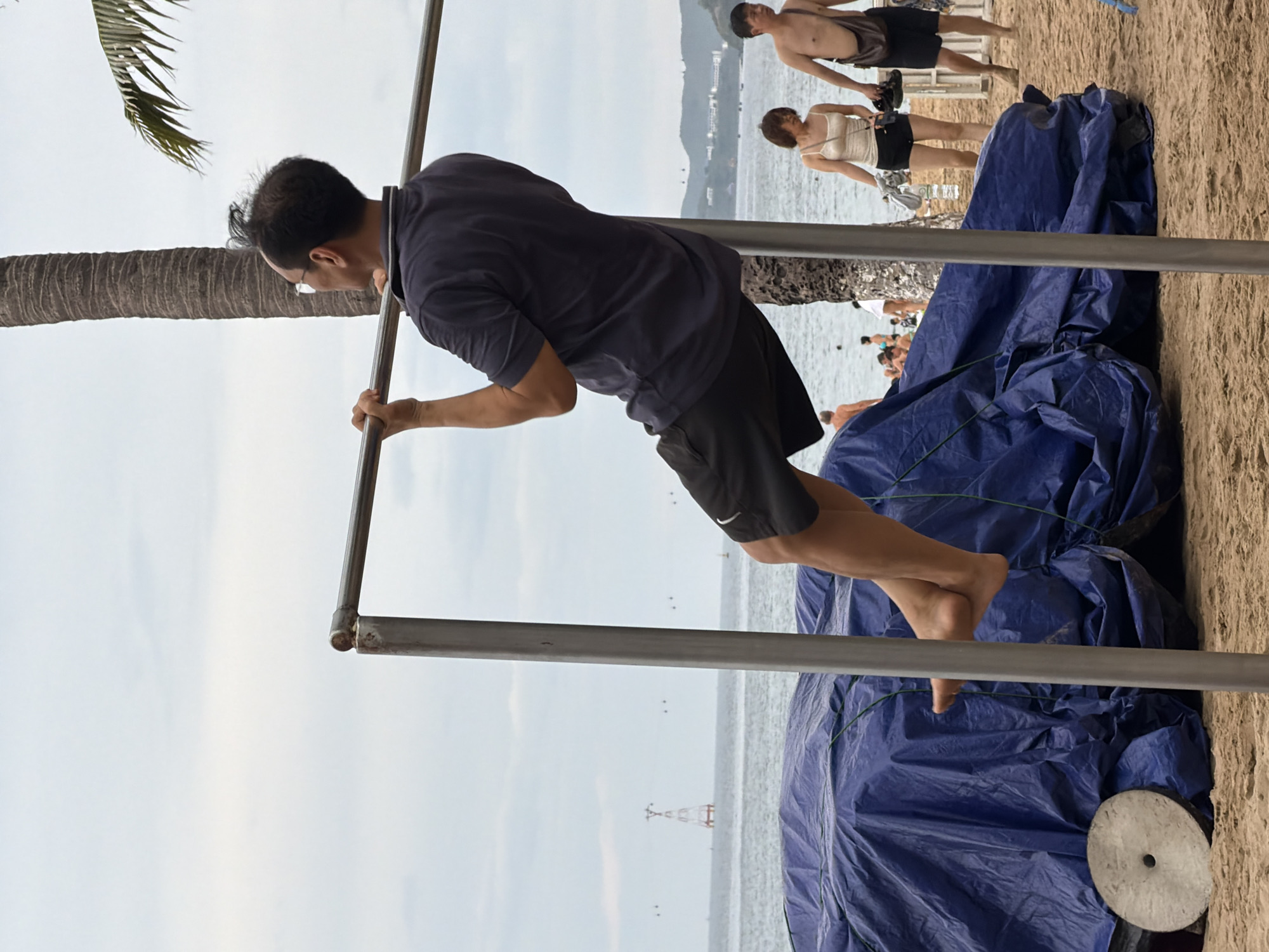 A man is doing pull-ups on a bar on a sandy beach in Nha Trang, Vietnam. He is wearing a dark blue t-shirt and black shorts. The ocean is visible in the background, with a few people on the beach and in the water. A large blue tarp covers some objects to the left of the pull-up bar. The sky is overcast.